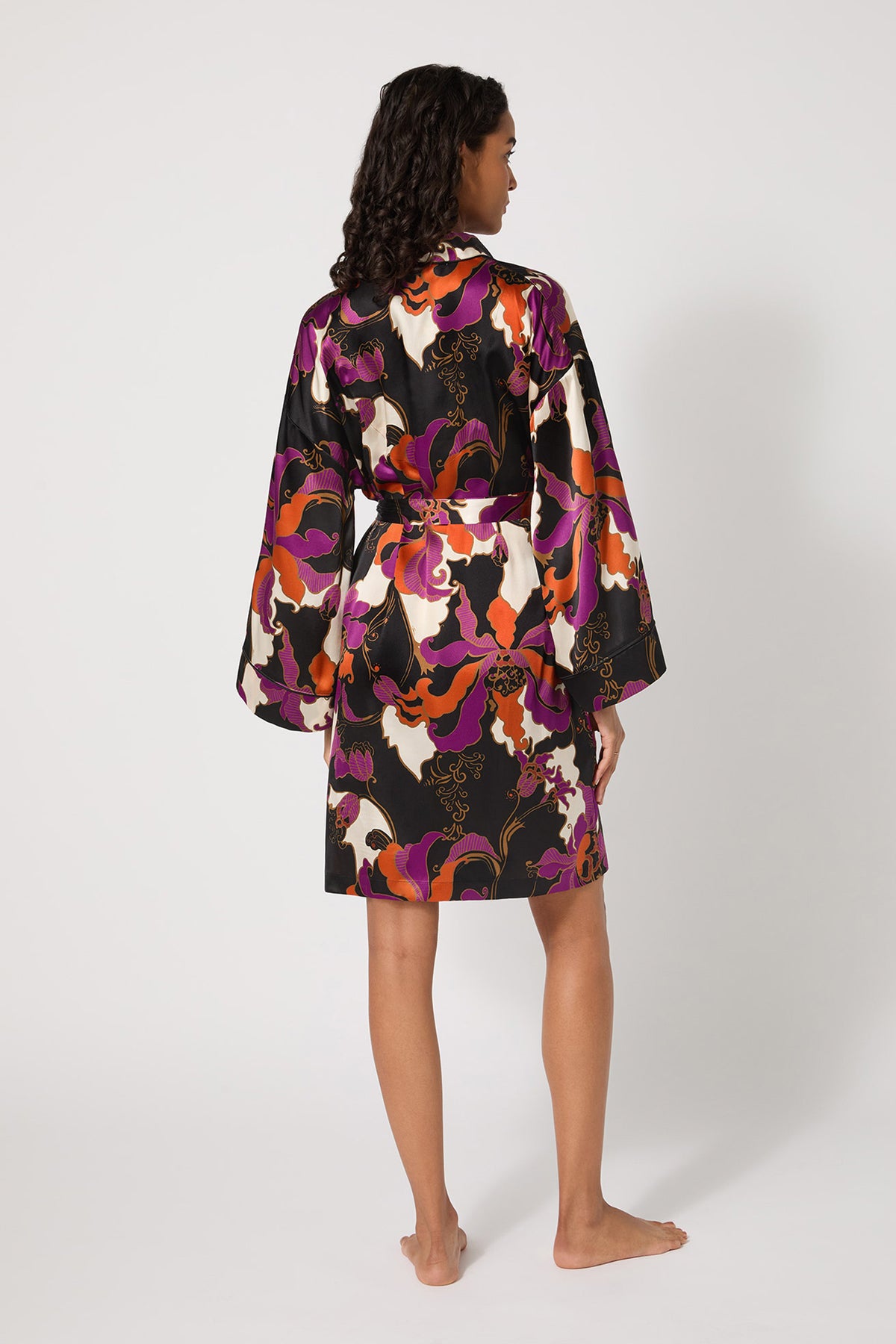A woman with curly hair stands barefoot, facing away, wearing the Long-Sleeve Silk Robe - Fall Floral featuring a bold purple, black, white, and orange floral pattern against a plain white background.