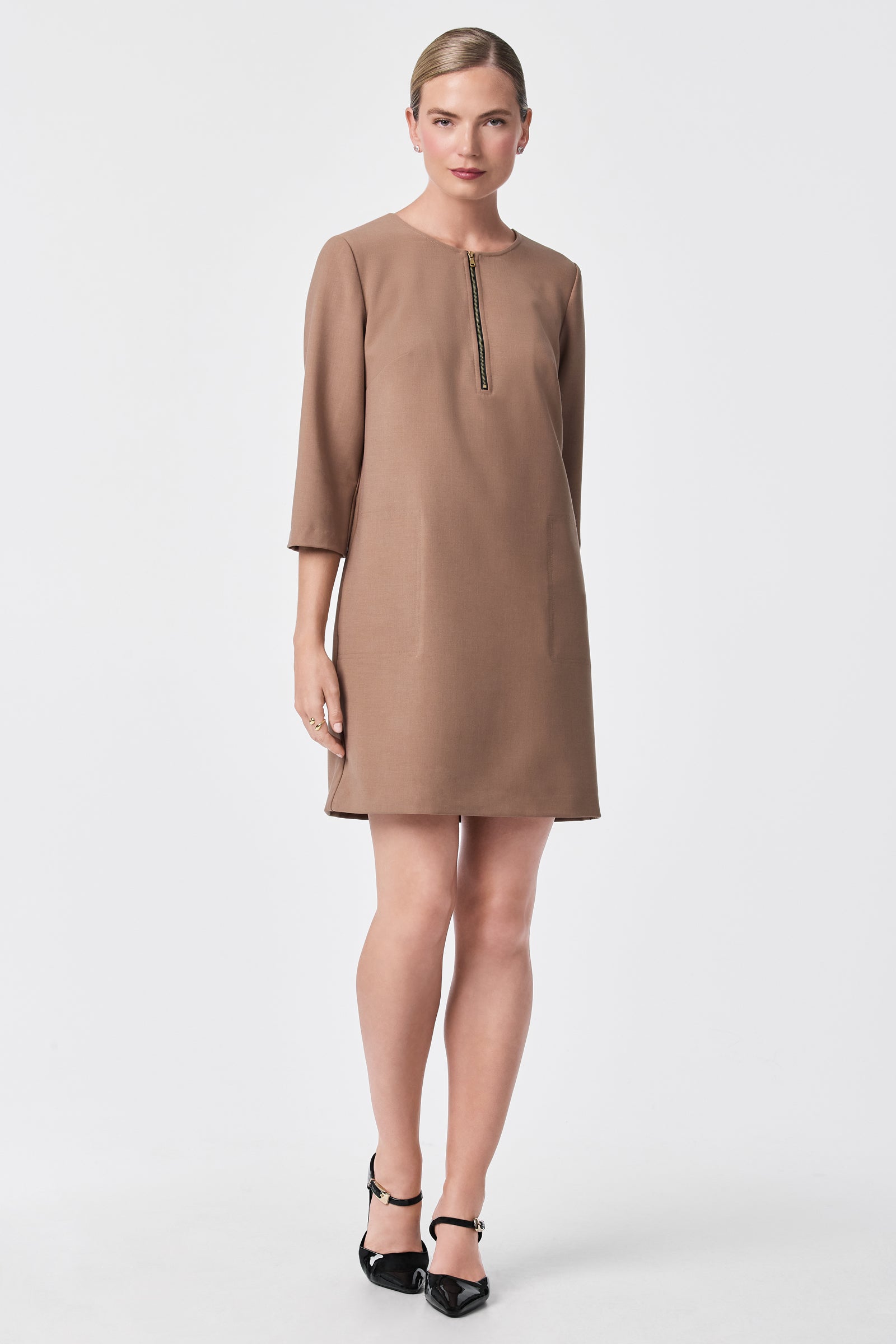 A woman stands against a plain white background wearing the Versed Dress—a knee-length, long-sleeved taupe dress with a front zipper detail. Her hair is pulled back and she completes the look with black heels.