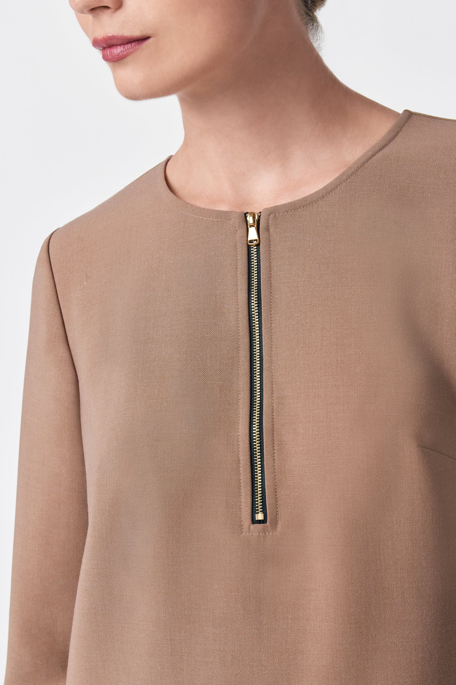 A woman shown from shoulders to chest wears the Versed Dress—a taupe long-sleeve top with a gold front zipper, partially unzipped, against a plain background.