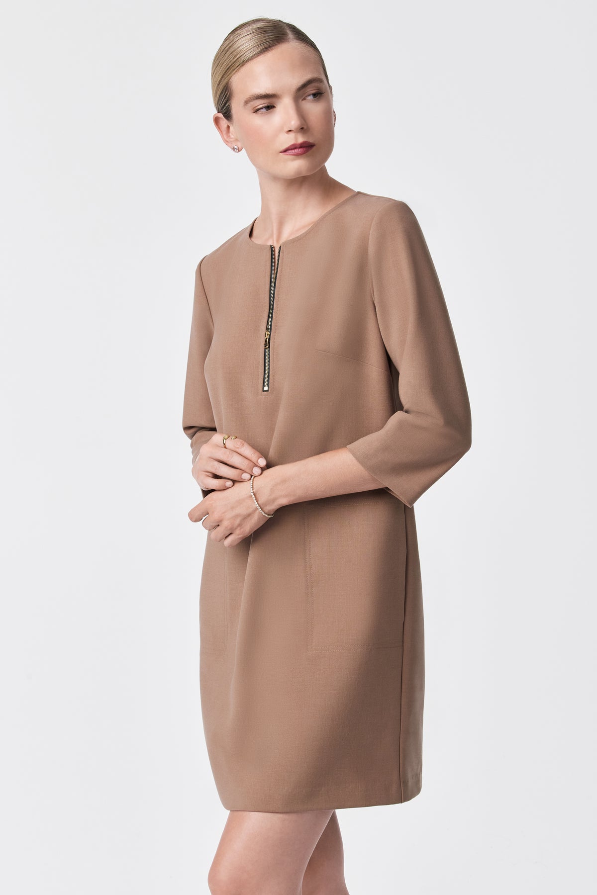 A woman models the Versed Dress—a tan, knee-length dress with three-quarter sleeves and a front zipper—standing against a white background, gazing left with her hands lightly clasped.