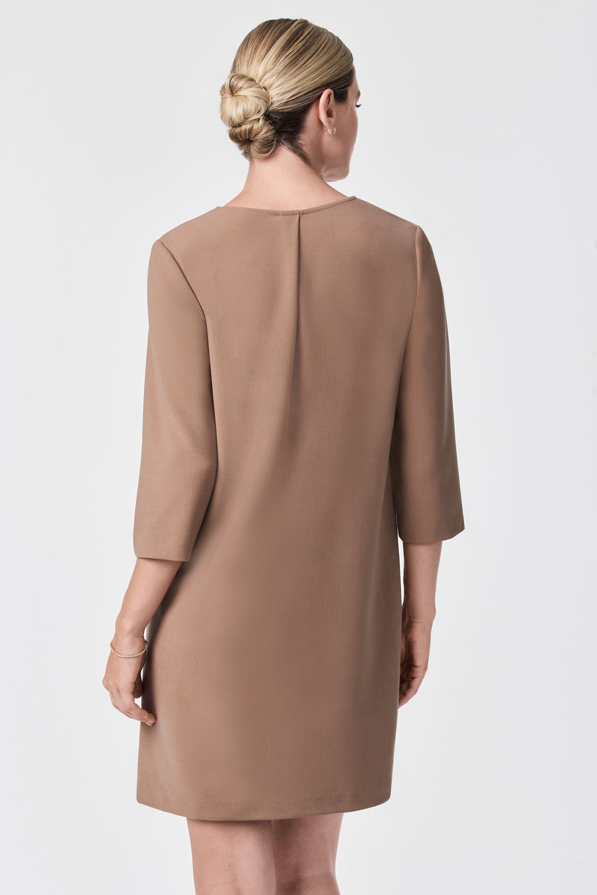 A woman with blonde hair in a neat low bun models the Versed Dress, a knee-length, camel-colored piece with three-quarter sleeves, shown from the back against a plain white background.