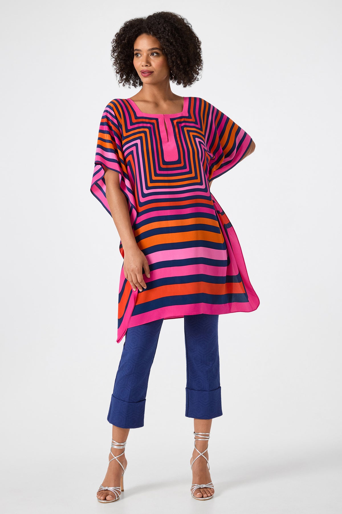 A woman with curly hair poses against a white background wearing Banshee Cropped Cuff Hem Pants, a colorful square-striped tunic in pink, orange, and navy, and silver strappy heels, with one hand on her hip.