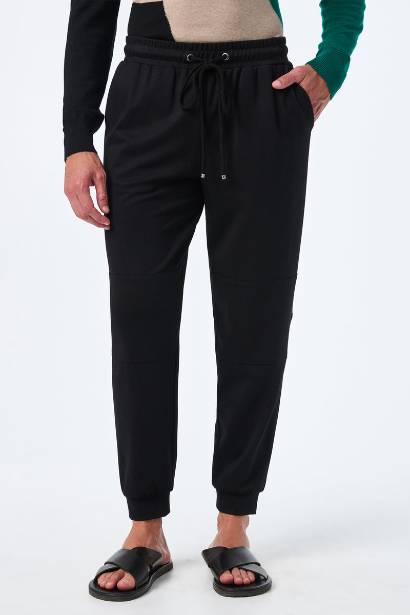 Someone wearing the Hideaway Jogger—black joggers with a drawstring waist and side pockets—paired with black slide sandals and a color-block sweater, stands against a white background.