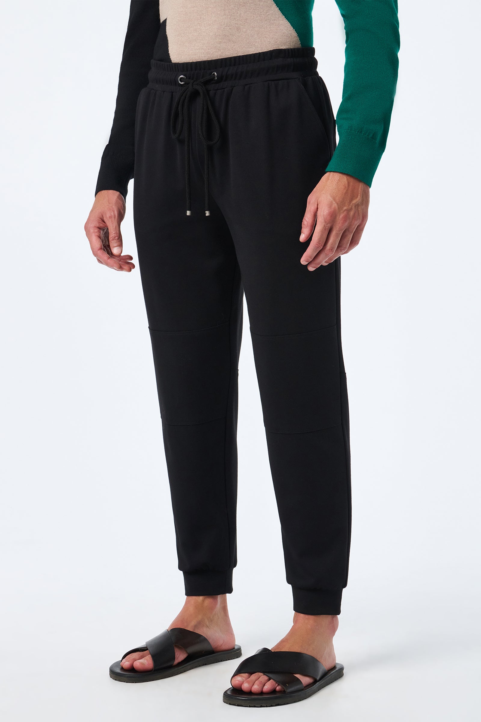A person in Hideaway Jogger pants with a drawstring waist and cuffed ankles, paired with a green and black long-sleeve top and black slide sandals, stands against a plain white background.