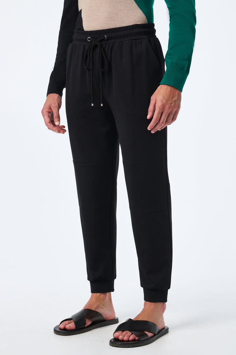 A person in Hideaway Jogger pants with a drawstring waist and cuffed ankles, paired with a green and black long-sleeve top and black slide sandals, stands against a plain white background.