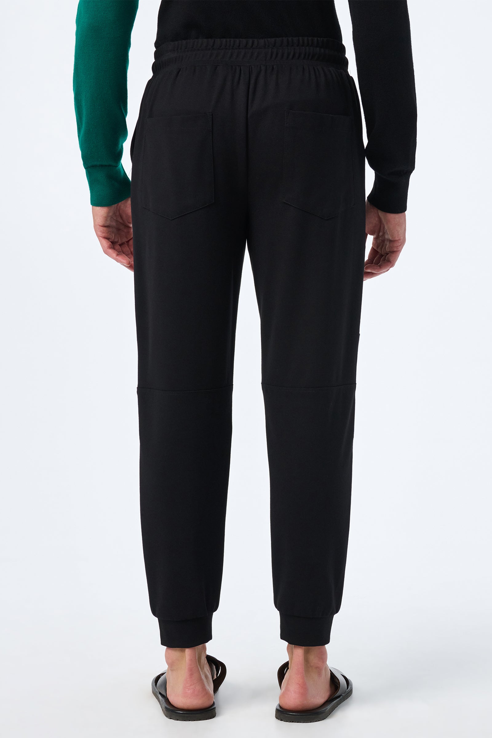 A person is shown from the back wearing Hideaway Jogger pants in black with black sandals and a green and black long-sleeve shirt, standing against a plain white background, hands at their sides.