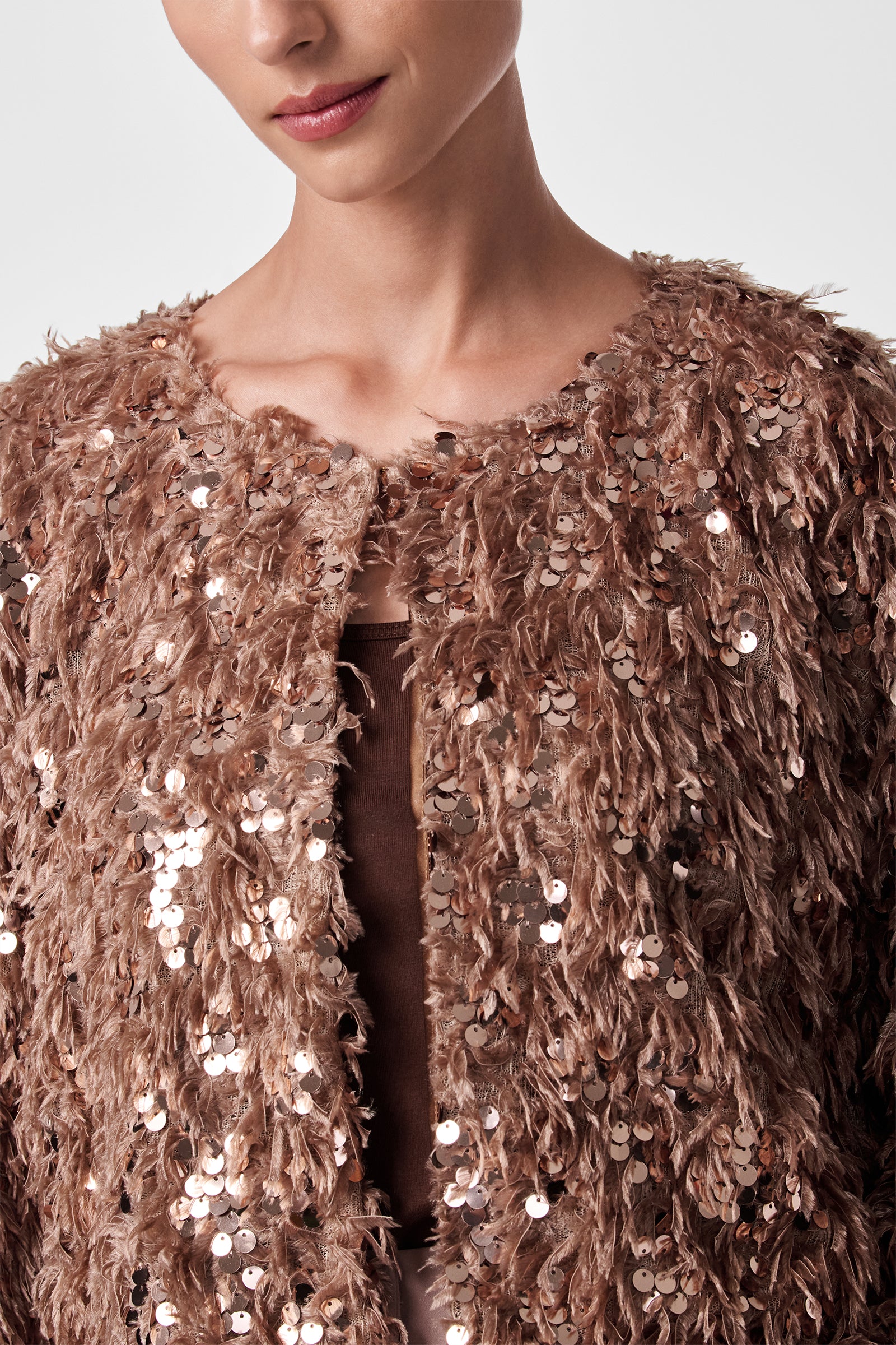 A woman wears the Fete Faux Fur Stole, a textured brown accessory with shiny sequins, over a brown top. She is shown from shoulders to mouth against a plain background.