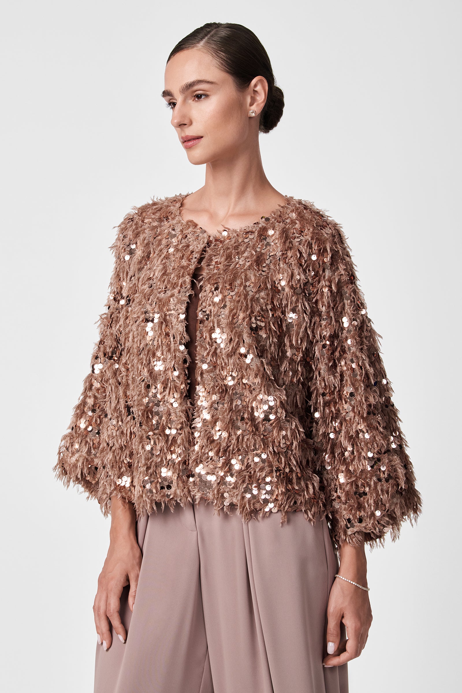 A woman with dark hair in a sleek bun wears the Fete Faux Fur Stole, styled over taupe pants, standing against a plain light background.