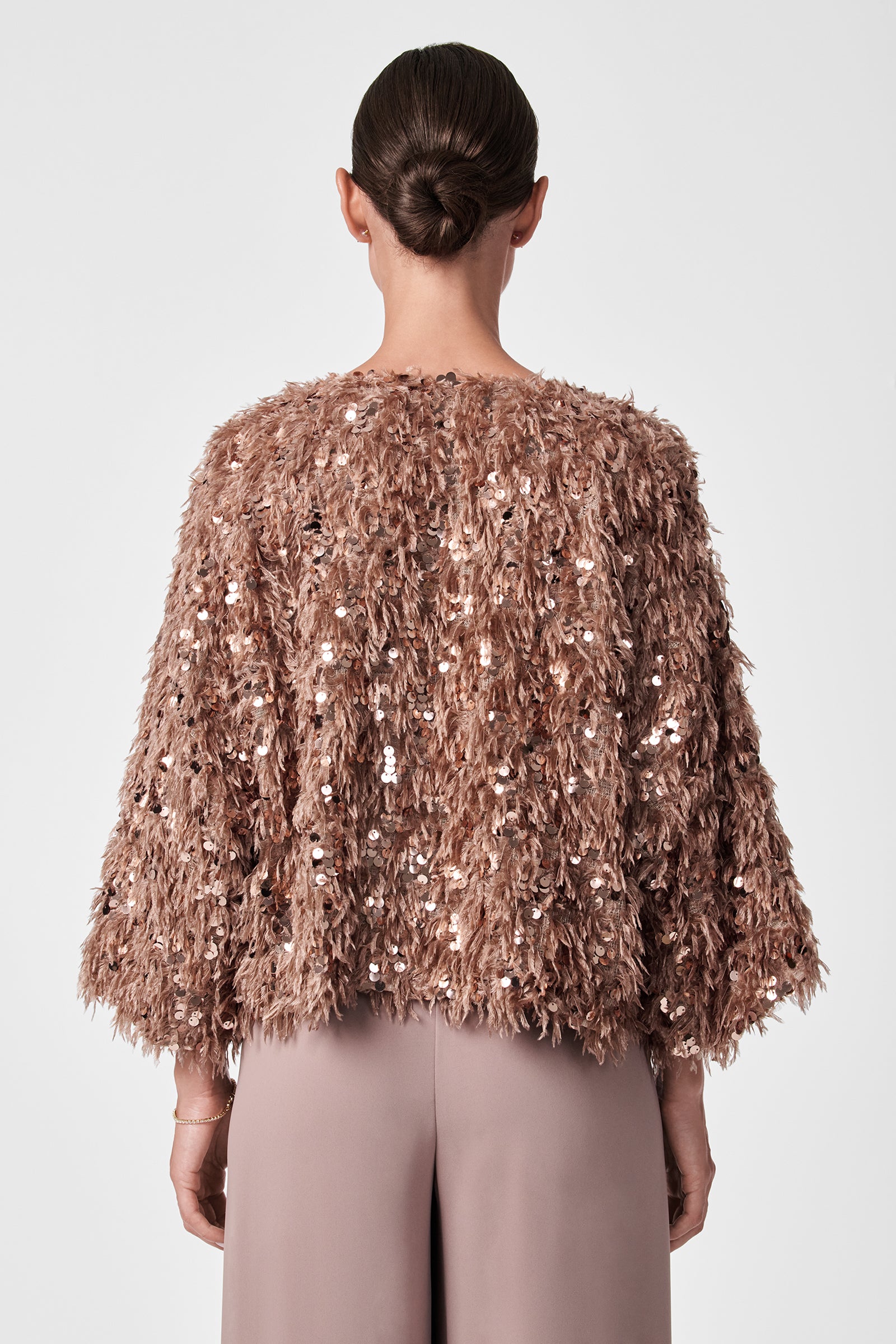 A woman with dark hair in a sleek bun is seen from behind, wearing the Fete Faux Fur Stole over loose, taupe pants against a plain background.