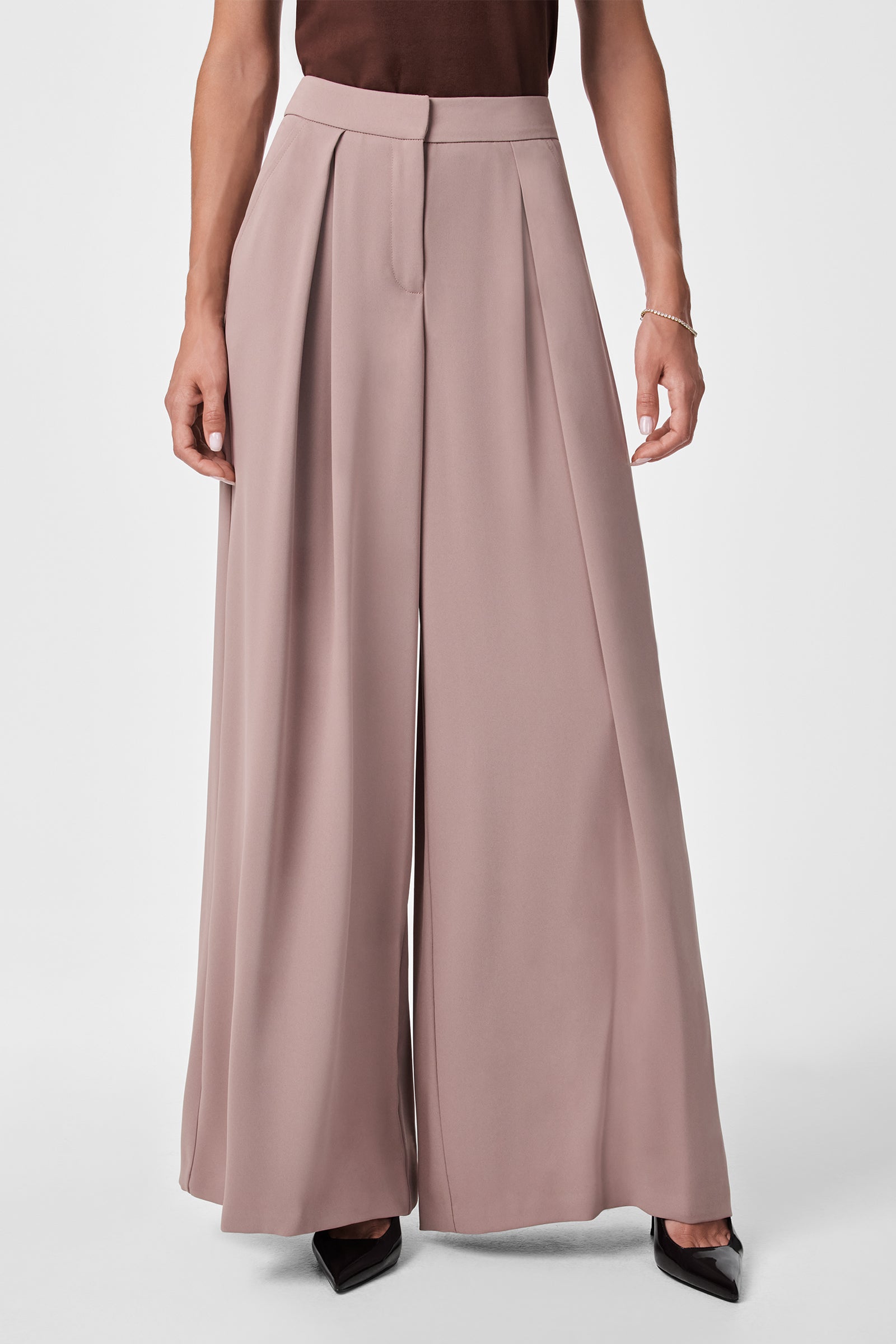 A person wearing Saige Wide-Leg Short Pants in taupe, featuring high waist, pleats, front zipper, and seam, paired with a dark top and black pointed-toe shoes. Only their lower torso and legs are visible.