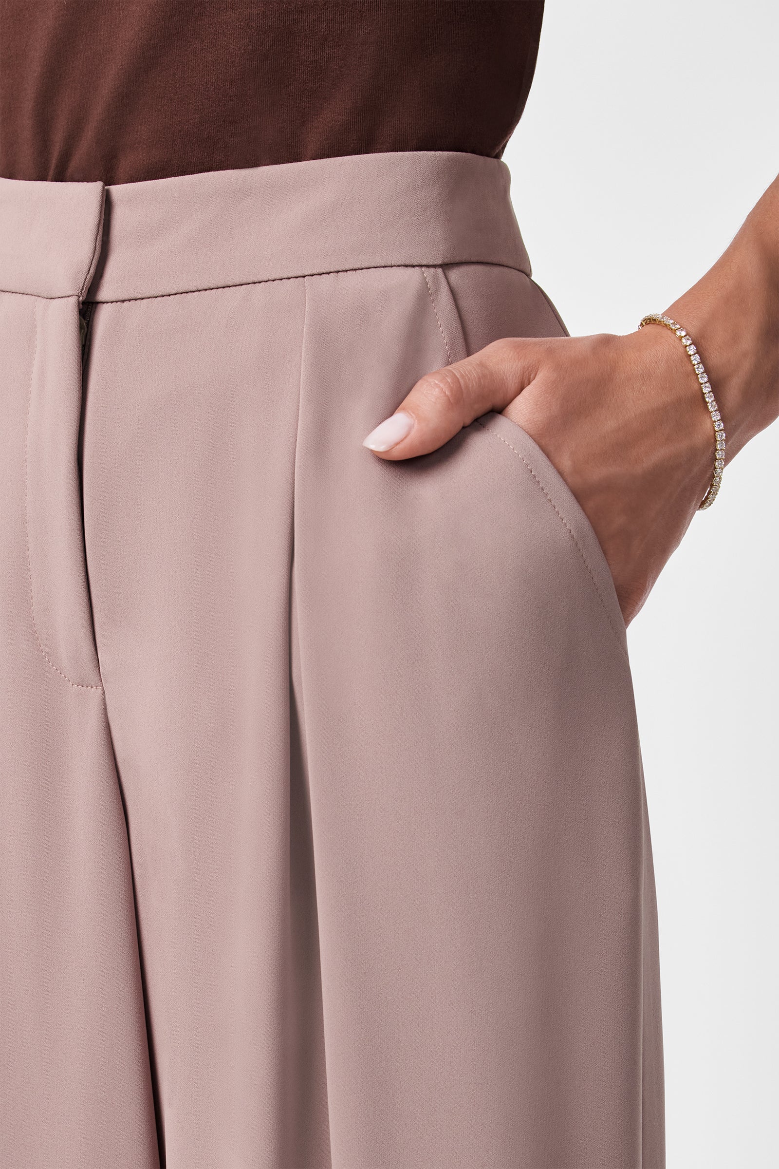 A person wears a brown top and Saige Wide-Leg Short Pants in light mauve, with their right hand in the pocket and a delicate gold bracelet on their wrist.