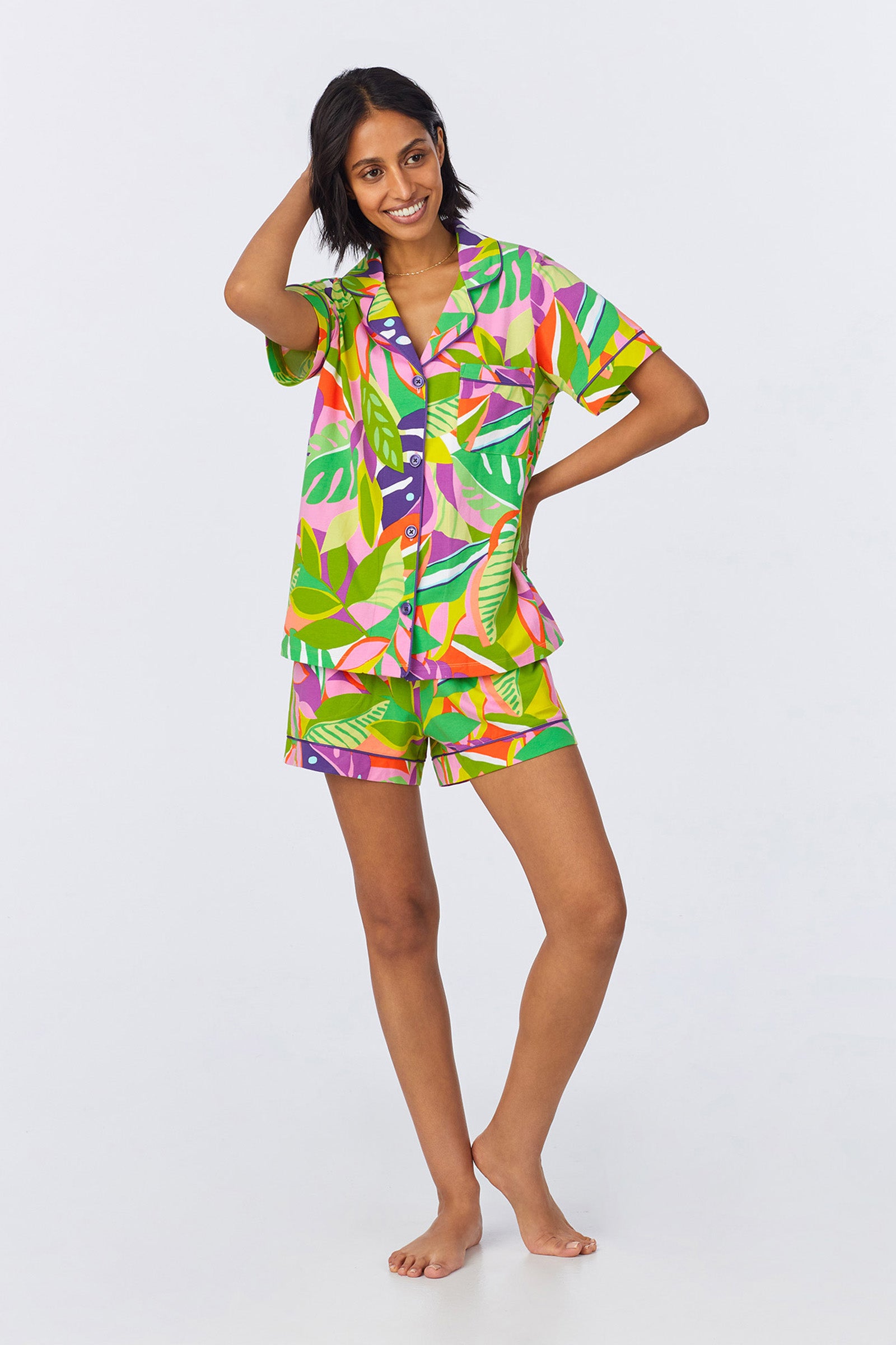 A woman stands barefoot, smiling, in the Shorty PJ Set - Jungle Stretch Jersey: a matching short-sleeve button-up shirt and shorts featuring a vibrant tropical leaf print in green, purple, pink, and yellow on a light background.