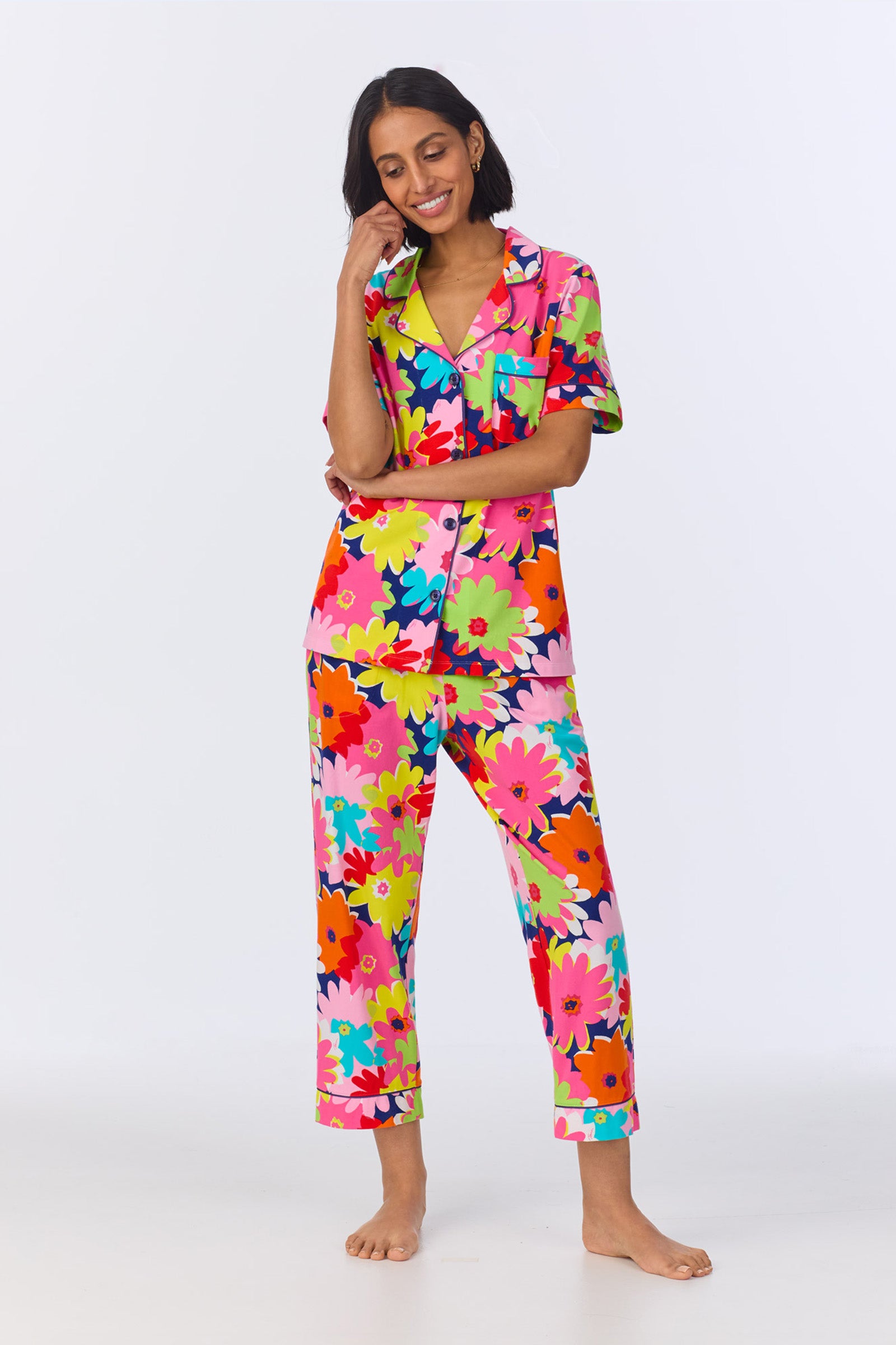 A woman stands barefoot, smiling, and wearing the Crop Classic PJ Set - Pop Floral Stretch Jersey in Multi, featuring a bold, multicolored floral top and pants against a plain white background.