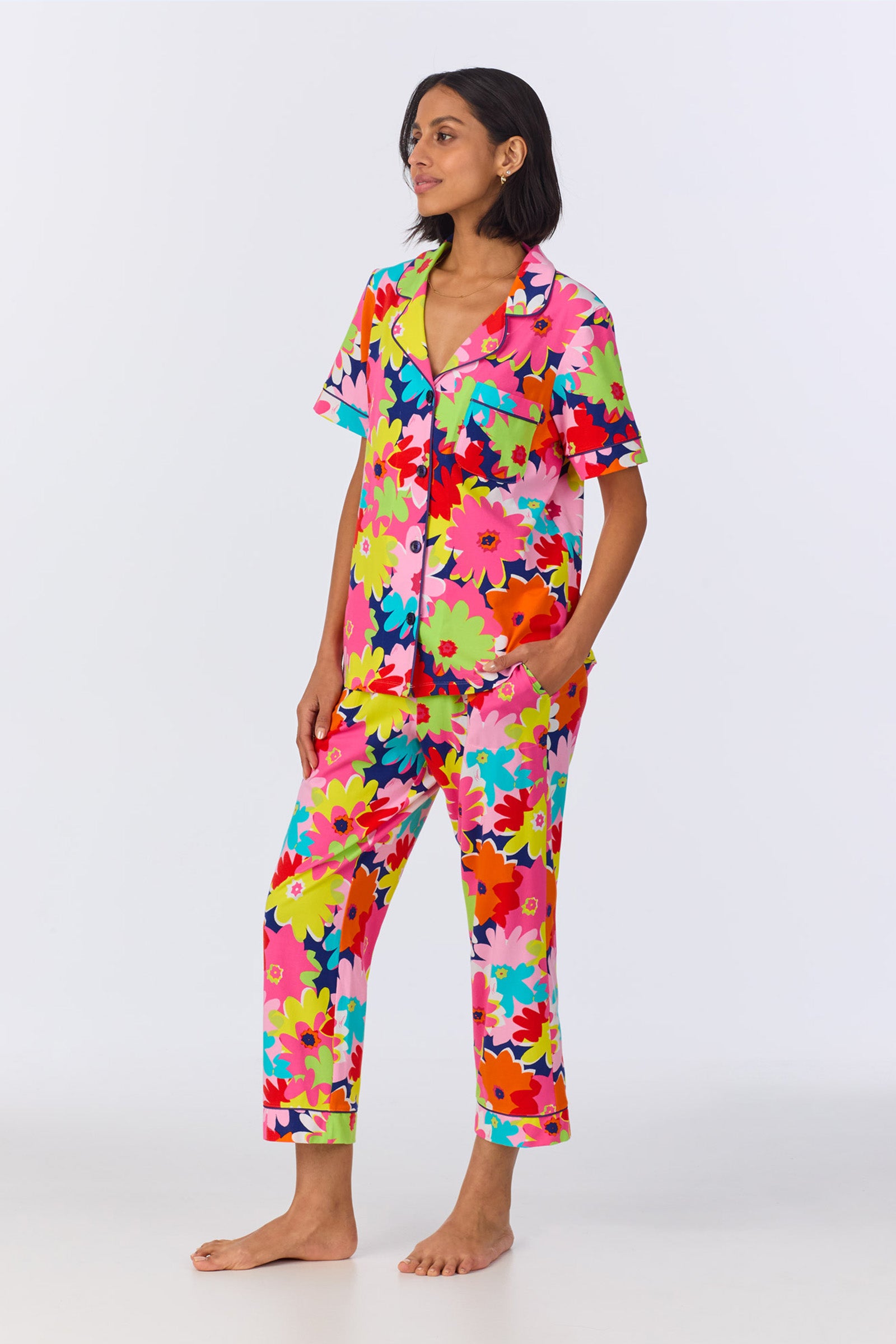 A woman stands barefoot wearing the Crop Classic PJ Set - Pop Floral Stretch Jersey, a colorful pajama set with short sleeves and long pants featuring bright pink, red, green, yellow, ochre, and blue flowers on a white background.