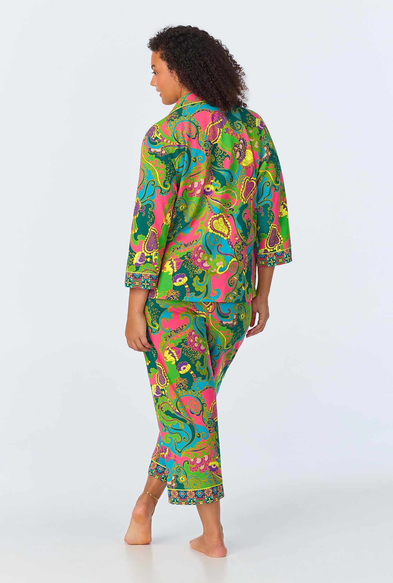 A person stands barefoot, facing away, wearing the Turkish Delight Womens 3/4-Sleeve Cropped Pant Jersey PJ Set—vibrant Trina Turk sleepwear with a green, pink, yellow, and blue paisley and floral pattern.