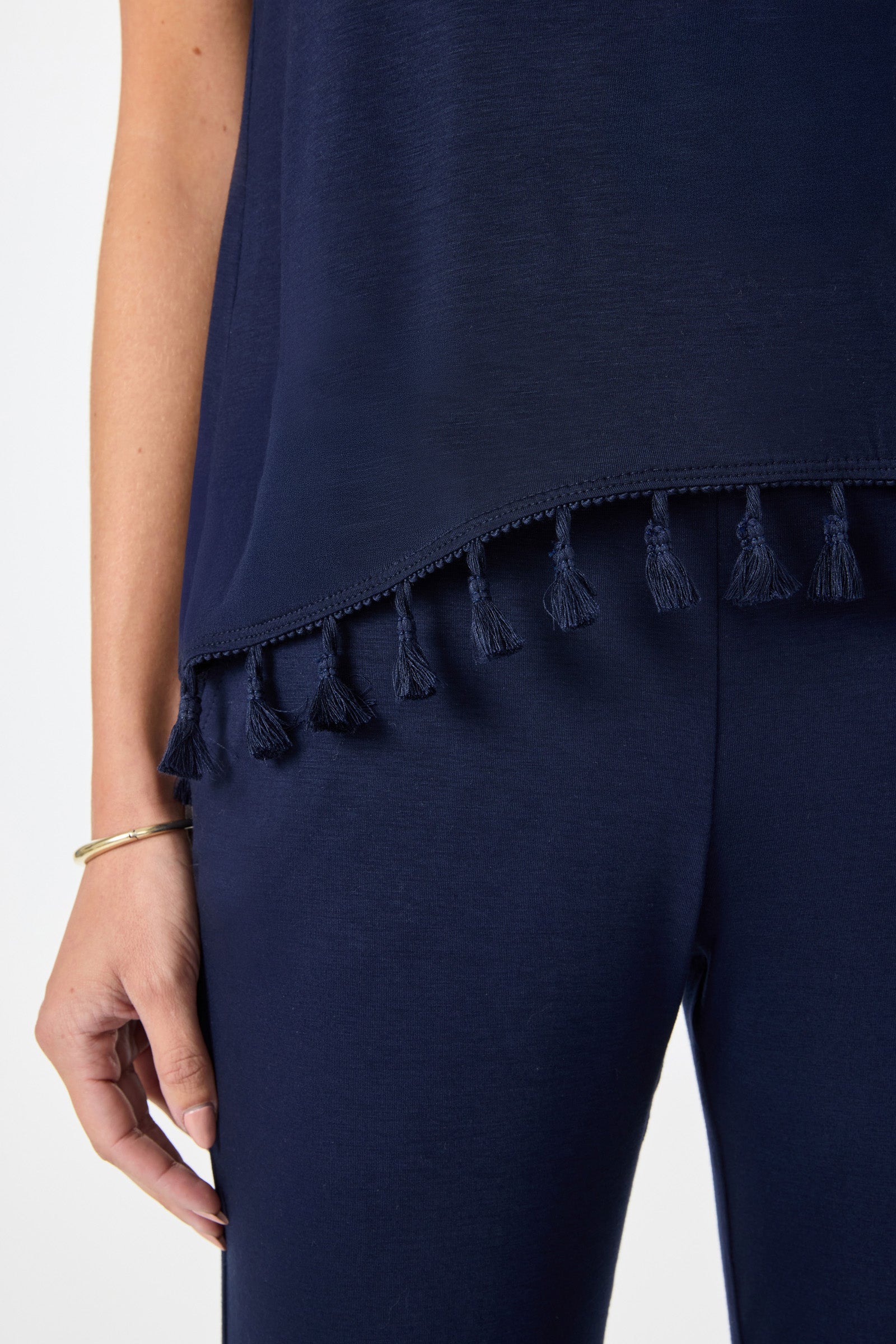 A person wears the Tajo 2 Tassel Hem Sleeveless Top in navy blue, paired with matching pants. Their left hand, adorned with a gold bangle and white nail polish, rests relaxed at their side.