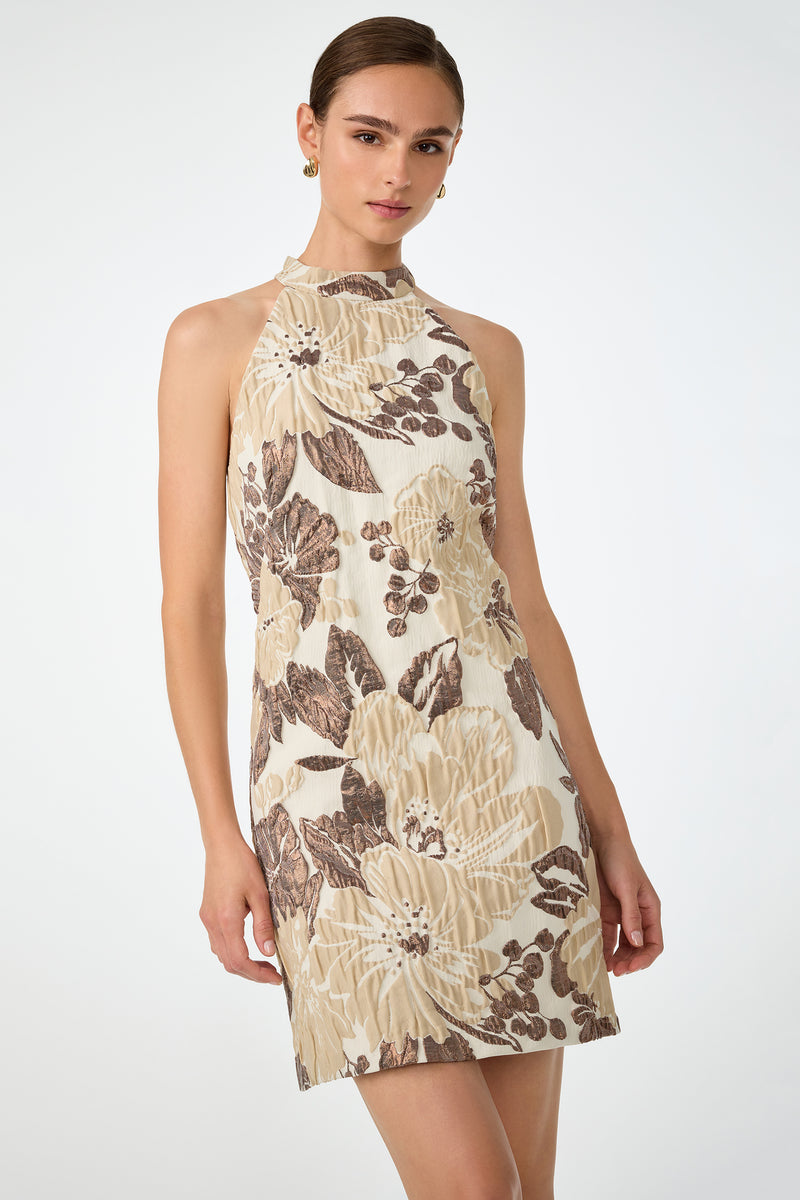 A woman models the Garam Sleeveless Halter Dress, featuring a high neck and beige mini silhouette with bold brown floral patterns, against a plain light background.