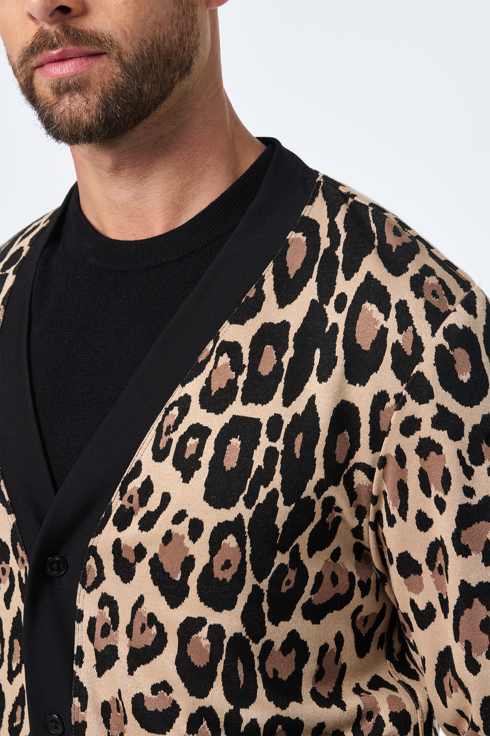 A man with a trimmed beard wears the Darryl Knit Cardigan, featuring a bold black and brown leopard print over beige, paired with a black shirt. The photo partially shows his face and upper body.