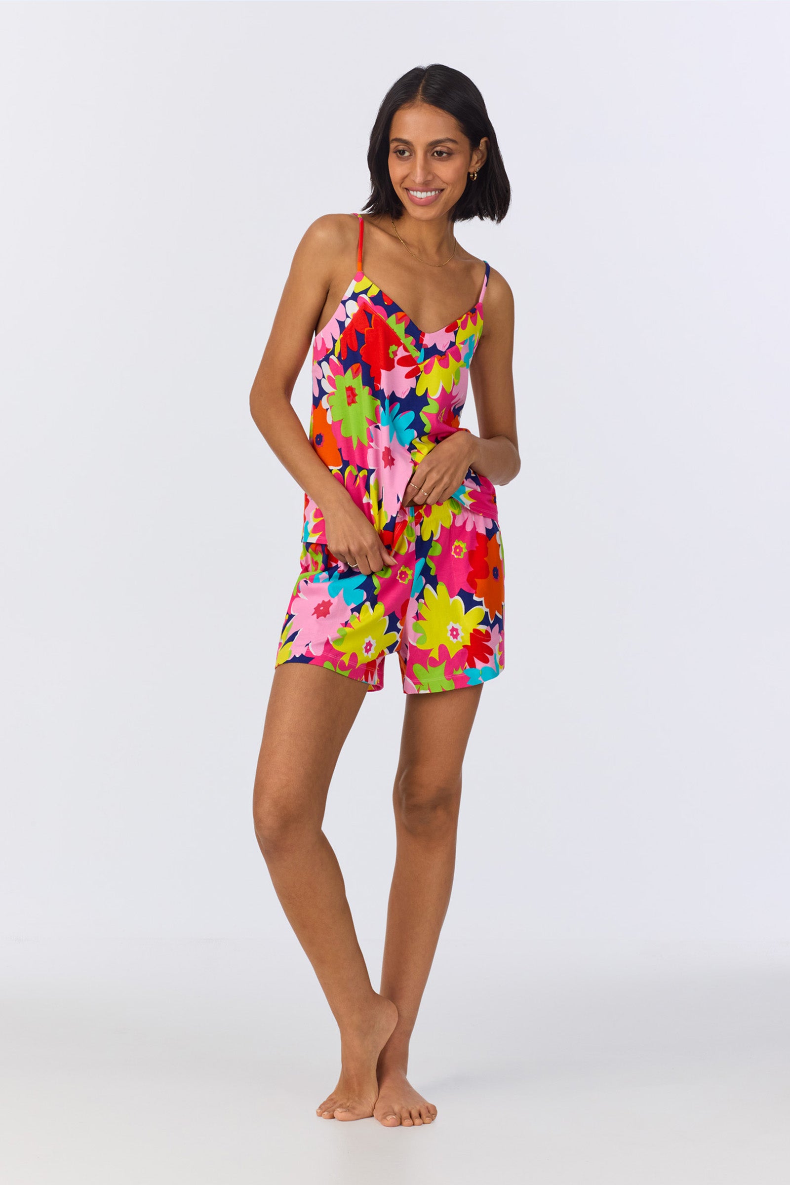 A woman stands barefoot, smiling and looking to the side, wearing the Cami Shorty PJ Set in multi Pop Floral Stretch Jersey against a plain white background.