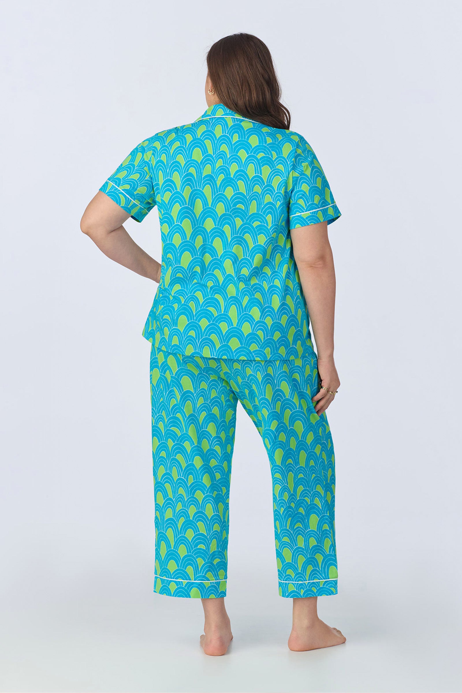 A person with long brown hair stands barefoot, facing away, wearing the Crop Classic PJ Set - Geo Stretch Jersey in blue and green with a repeating scallop pattern, against a plain light background.