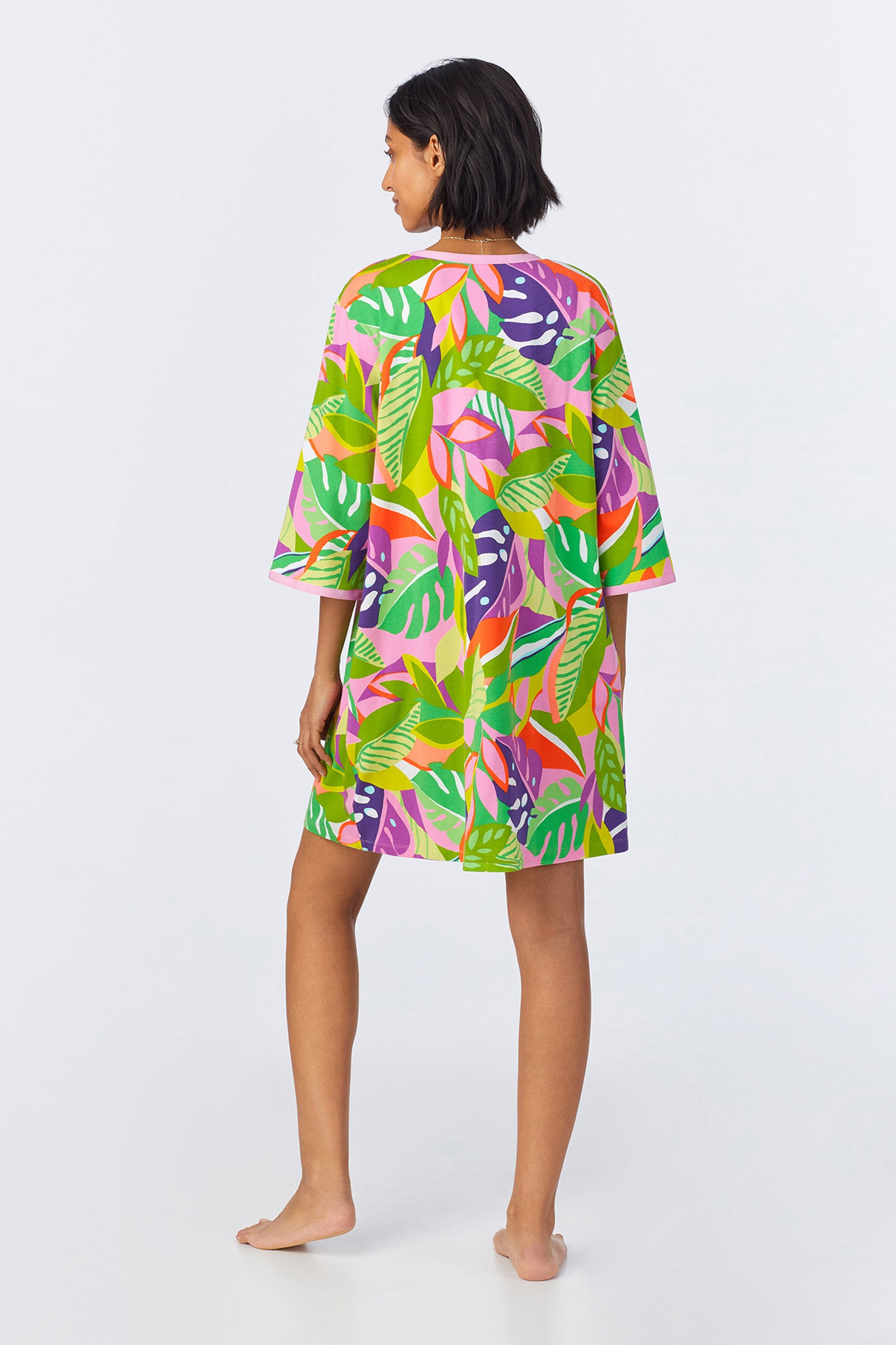 A woman stands barefoot, facing away, wearing the PJ Caftan Dress - Jungle Stretch Jersey. The dress features a loose fit and a vibrant tropical leaf pattern in green, pink, orange, and purple against a plain white background.