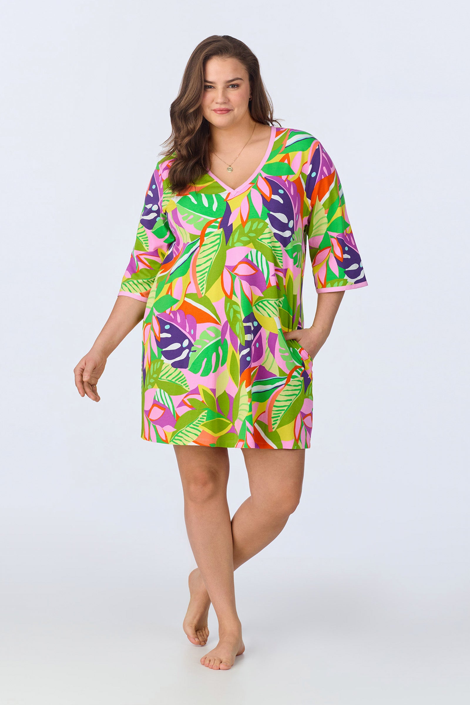 A woman with long brown hair smiles gently while wearing the PJ Caftan Dress - Jungle Stretch Jersey, a colorful tropical-patterned V-neck dress with three-quarter sleeves, standing barefoot against a plain light background.