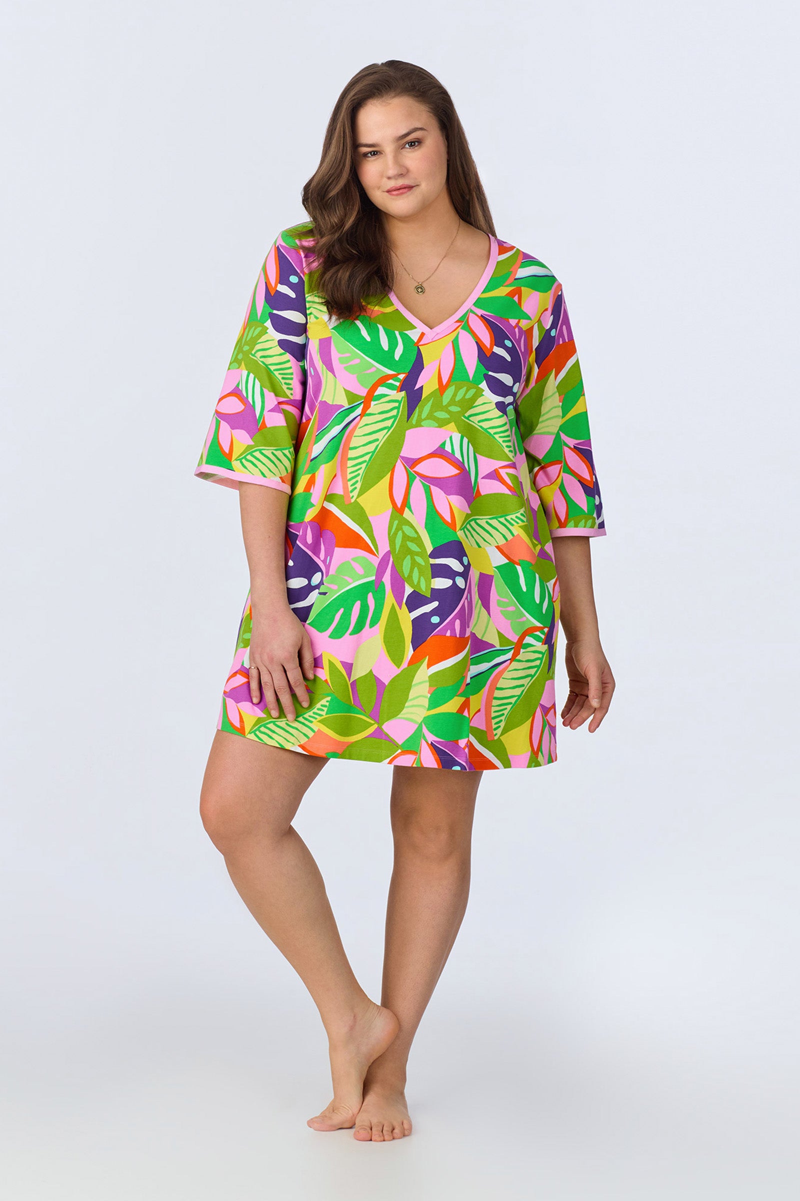 A woman with long brown hair confidently models the PJ Caftan Dress - Jungle Stretch Jersey, featuring vivid green, pink, yellow, and purple leaf prints. She stands barefoot against a plain light background, looking at the camera.