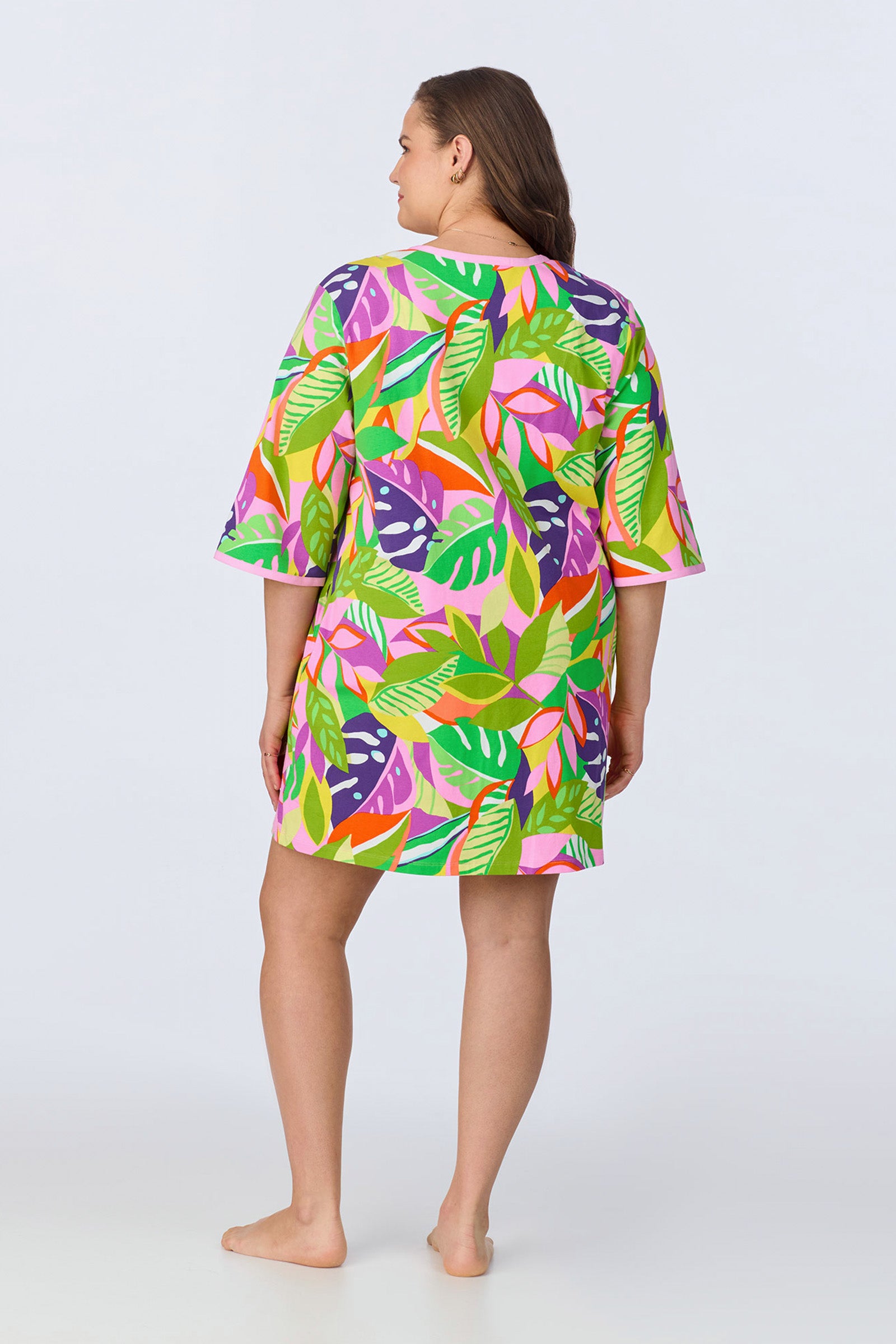 A woman stands barefoot, facing away, wearing the PJ Caftan Dress - Jungle Stretch Jersey, a knee-length dress with elbow-length sleeves and a vibrant green, pink, orange, and purple leaf pattern on a white background.