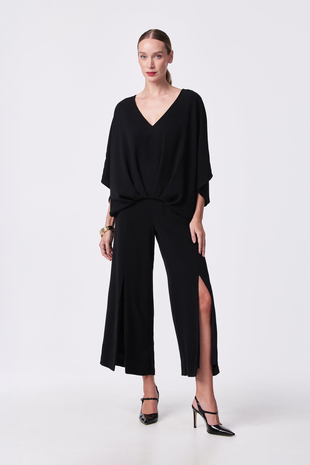A woman poses against a plain background in the Coralline 2 Top—a loose-fitting black V-neck—paired with wide-leg, high-slit black pants, black heels, gold bracelets, and her hair pulled back.