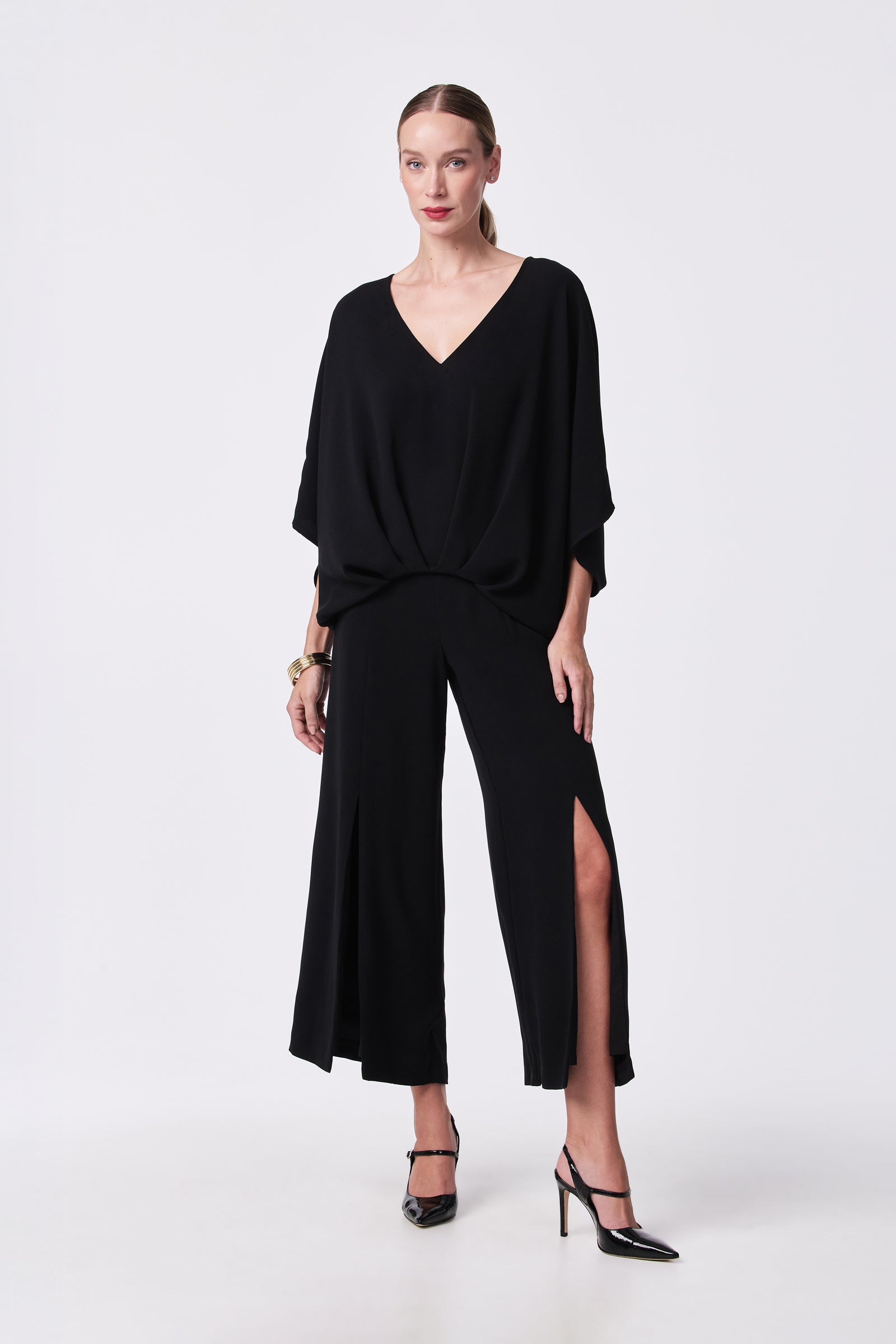 A woman poses against a plain background in the Coralline 2 Top—a loose-fitting black V-neck—paired with wide-leg, high-slit black pants, black heels, gold bracelets, and her hair pulled back.