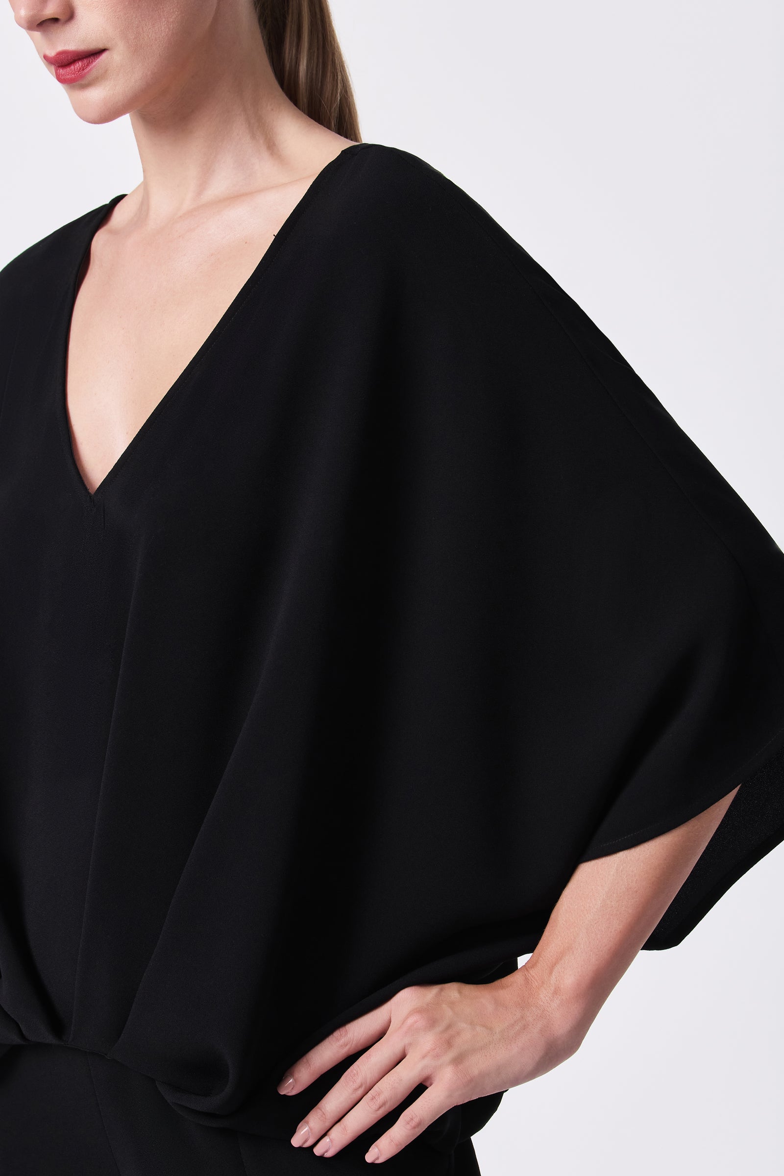 A woman models the Coralline 2 Top, a loose-fitting, elegant black blouse with wide batwing sleeves and a deep V-neckline, standing with one hand on her hip against a white background.