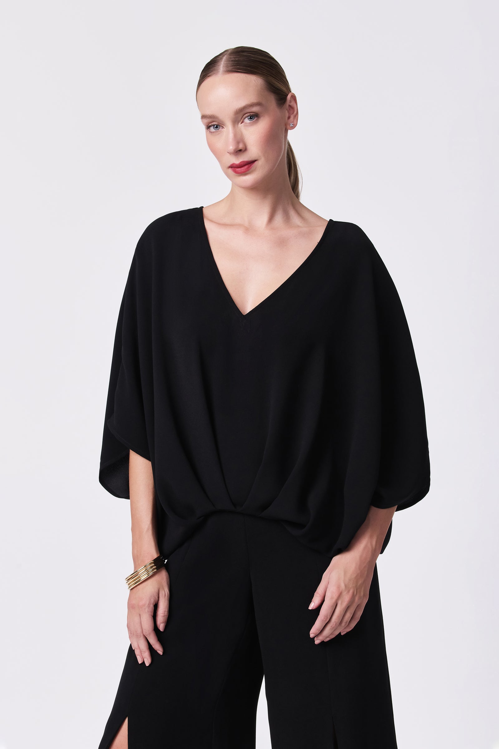 A woman with straight, light brown hair tied back wears the Coralline 2 Top, a loose-fitting black V-neck style, along with matching black pants. She stands against a plain white background, looking at the camera with a relaxed expression.