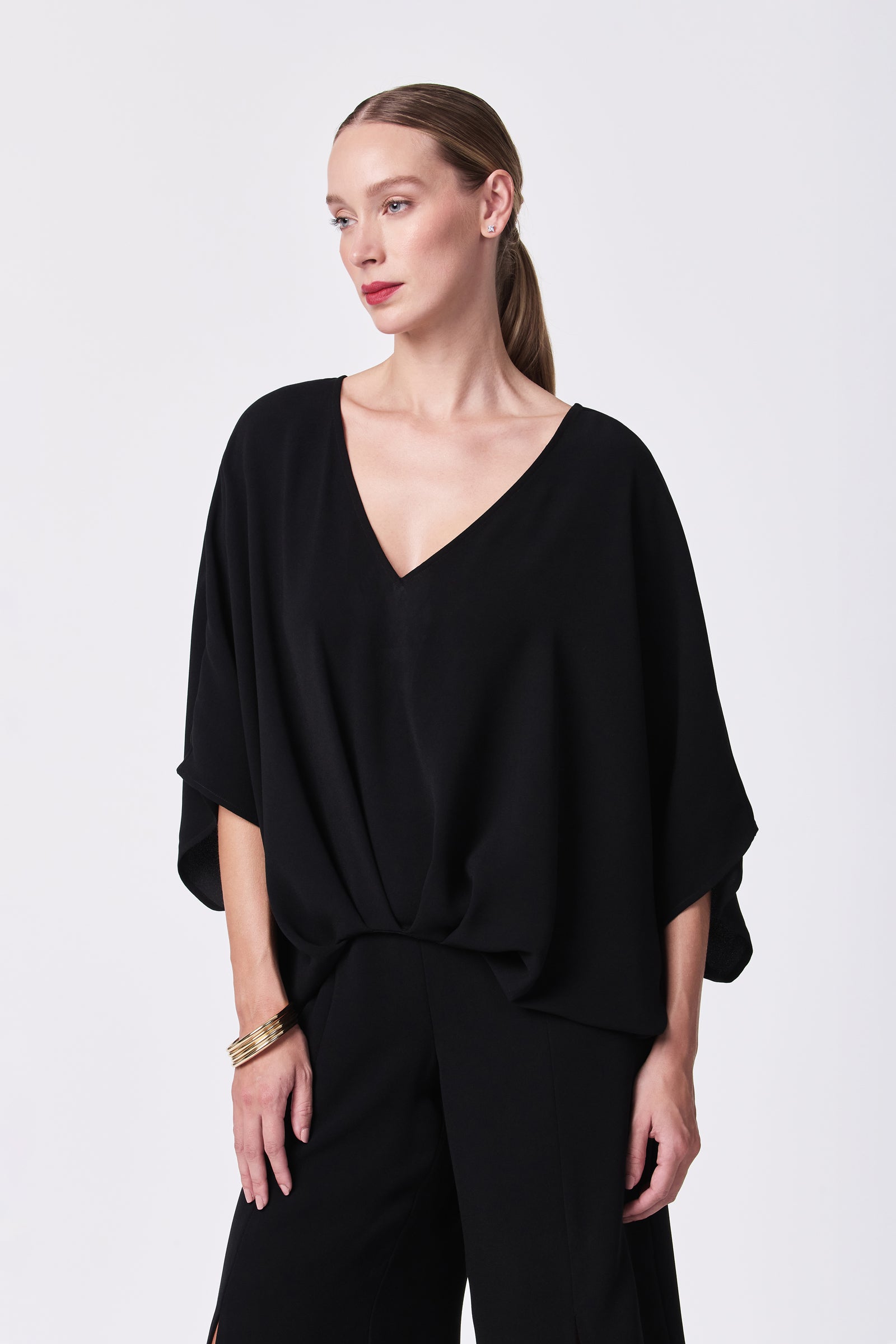 A woman with straight, light brown hair tied back wears the Coralline 2 Top—a loose, black V-neck blouse—paired with matching black pants and gold bangles, standing against a plain white background with a neutral expression.