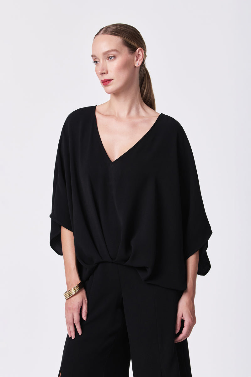 A woman with straight, light brown hair tied back wears the Coralline 2 Top—a loose, black V-neck blouse—paired with matching black pants and gold bangles, standing against a plain white background with a neutral expression.