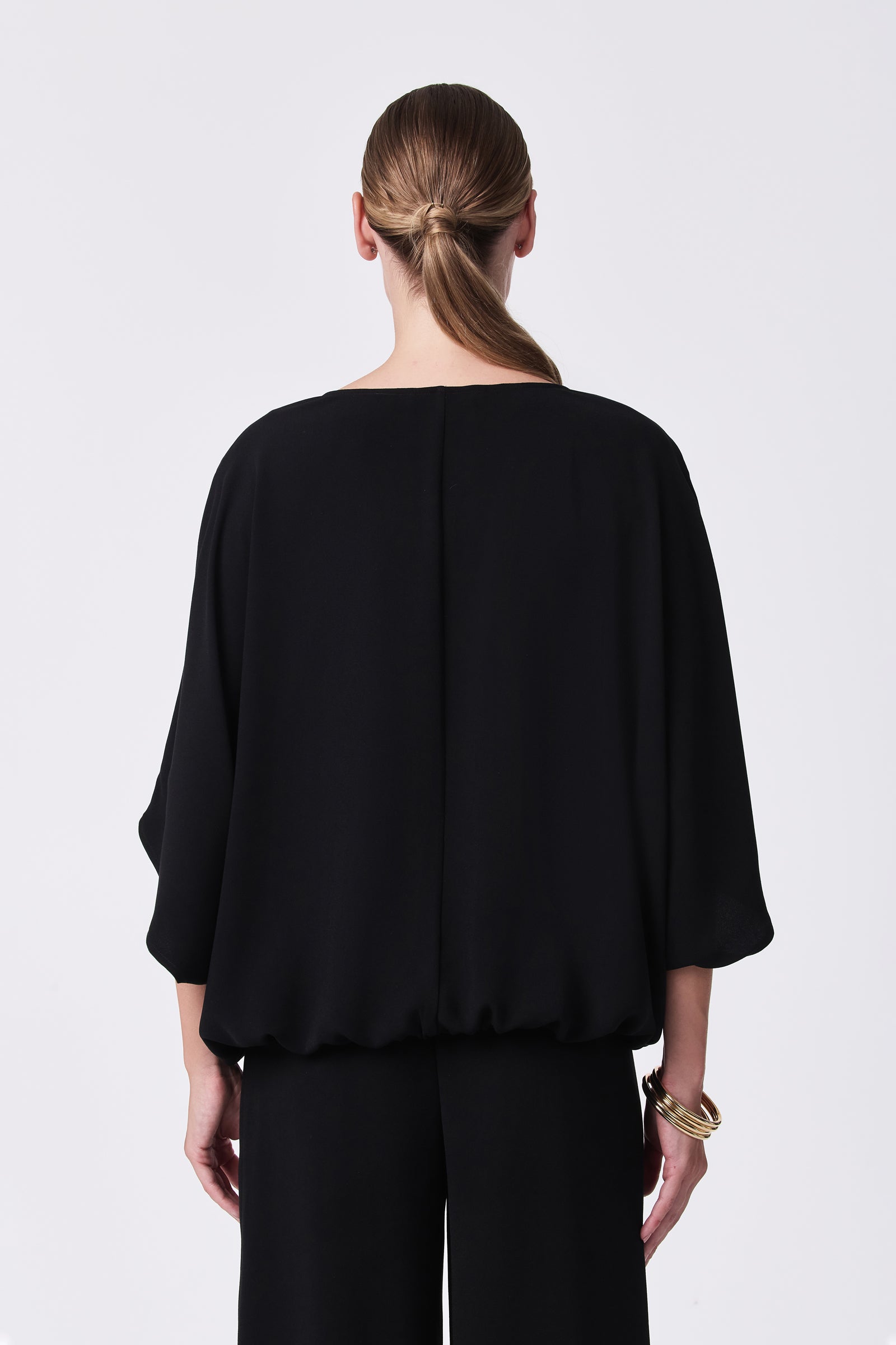 A woman with straight, light brown hair in a low knot stands facing away, wearing the loose-fitting Coralline 2 Top in black with wide sleeves and black pants. She accessorizes with gold bangles on her right wrist against a white background.