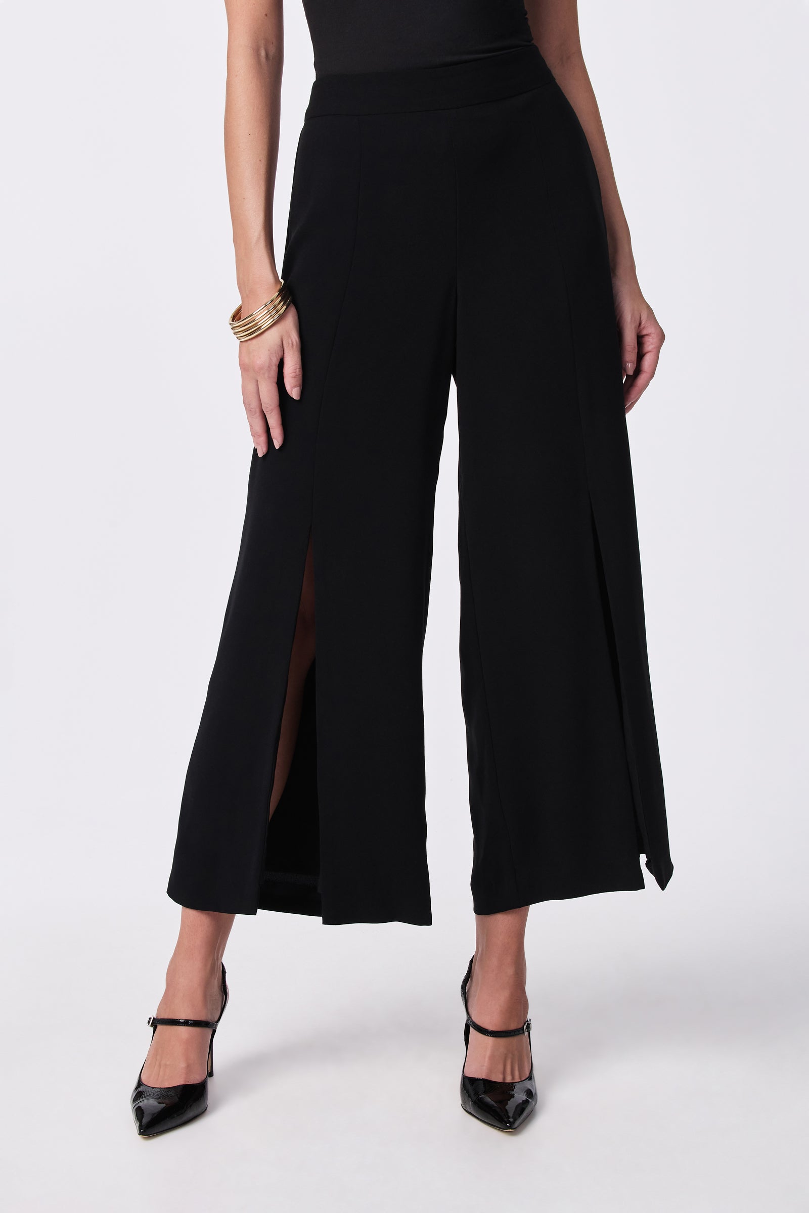 A person models the Davon Pant—wide-leg black pants with front slits—paired with a black sleeveless top, black heels, and gold bangles, standing against a plain light background.