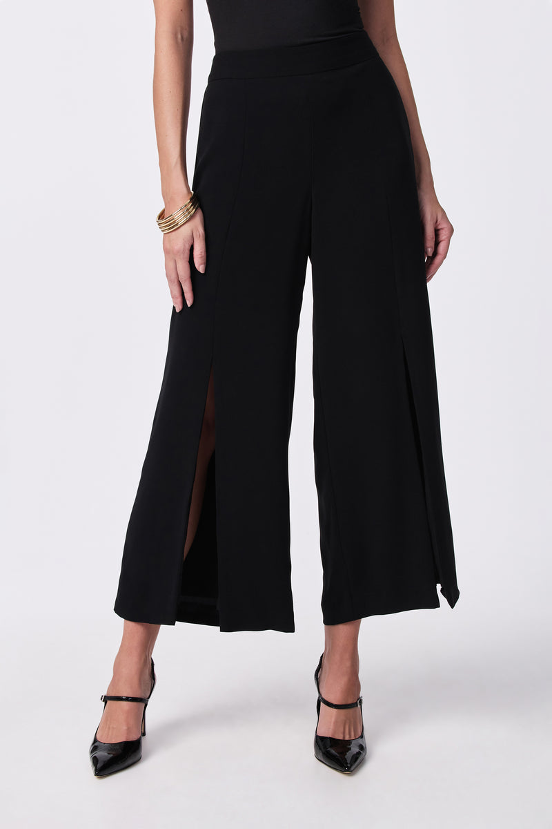 A person models the Davon Pant—wide-leg black pants with front slits—paired with a black sleeveless top, black heels, and gold bangles, standing against a plain light background.