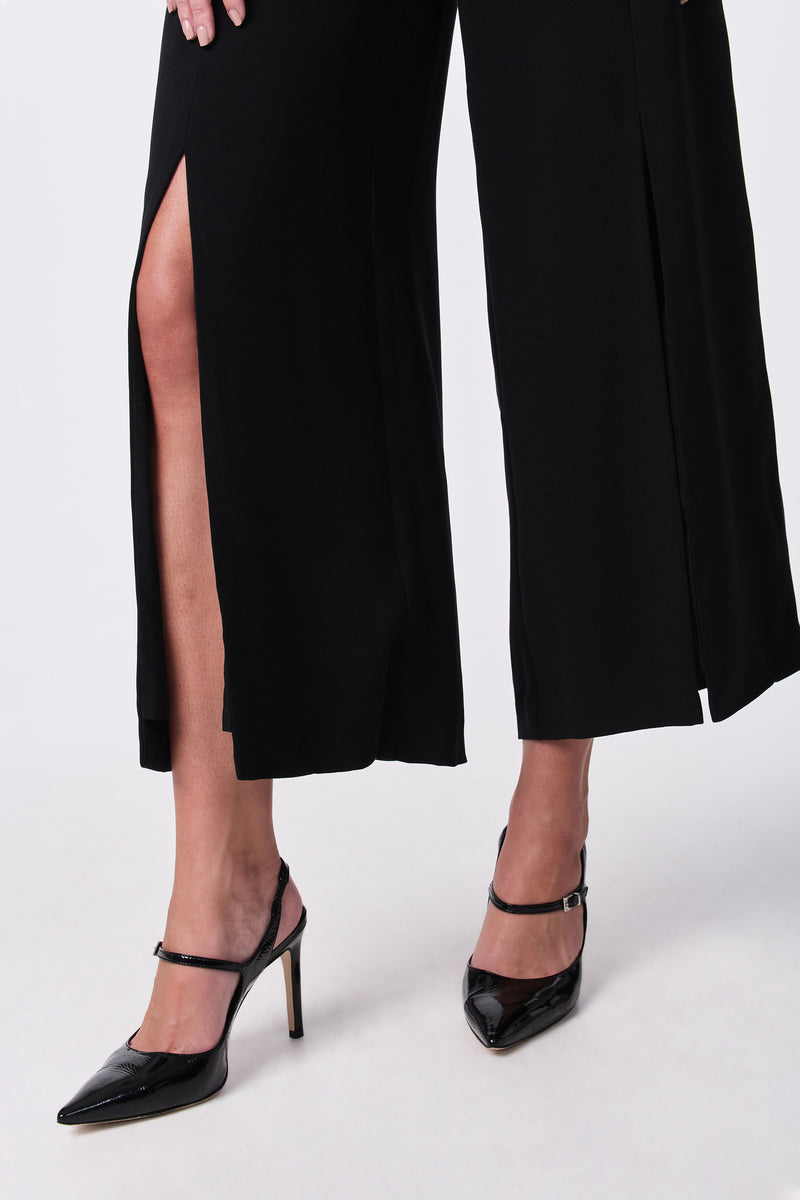 A person wearing the Davon Pant, black wide-leg pants with high slits, and black pointed-toe ankle-strap heels, standing against a plain white background.
