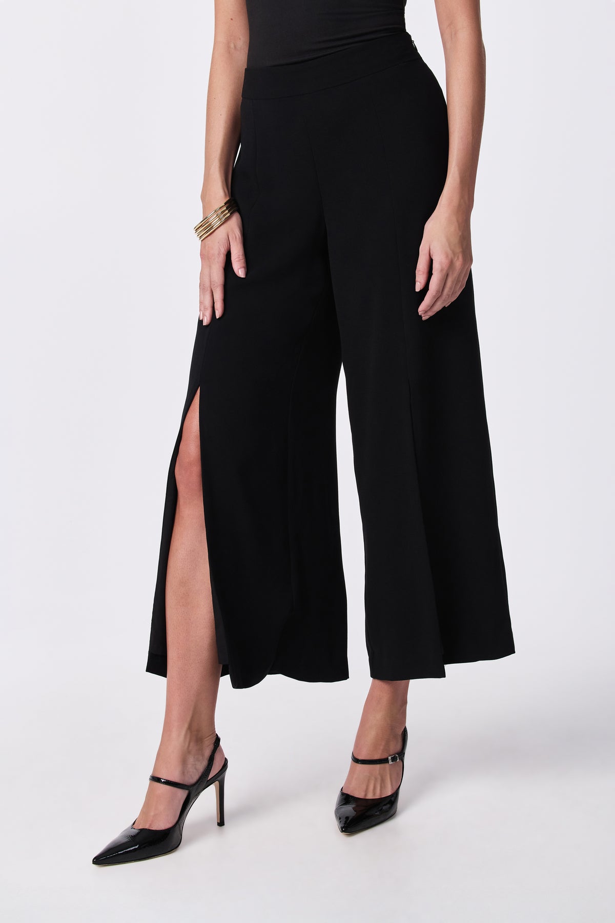 A person wearing the Davon Pant—black wide-leg culottes with a front slit—paired with black high heels, a black top, and gold bracelets, stands against a white background.