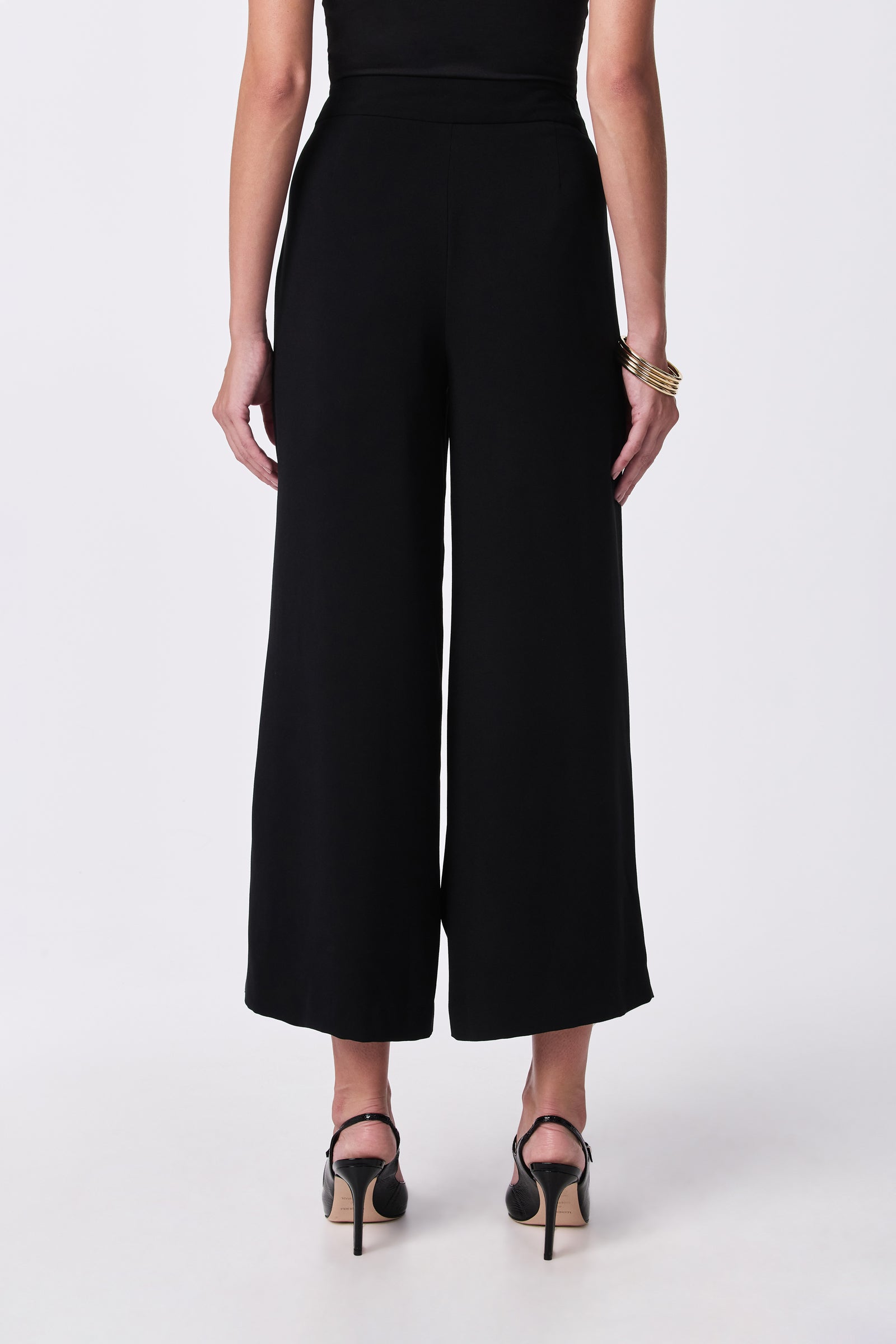 A woman faces away from the camera wearing the Davon Pant, a black top, black high-heeled sandals, and gold bangles on her left wrist, all set against a plain white background.