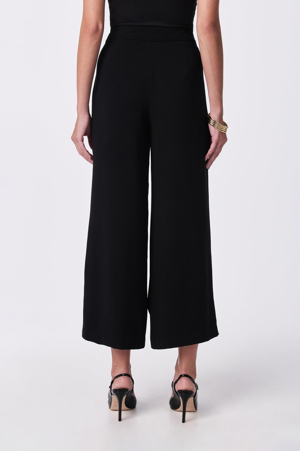 A woman faces away from the camera wearing the Davon Pant, a black top, black high-heeled sandals, and gold bangles on her left wrist, all set against a plain white background.