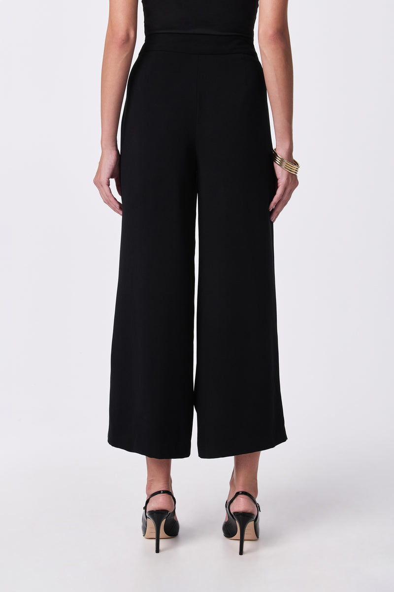 A woman faces away from the camera wearing the Davon Pant, a black top, black high-heeled sandals, and gold bangles on her left wrist, all set against a plain white background.