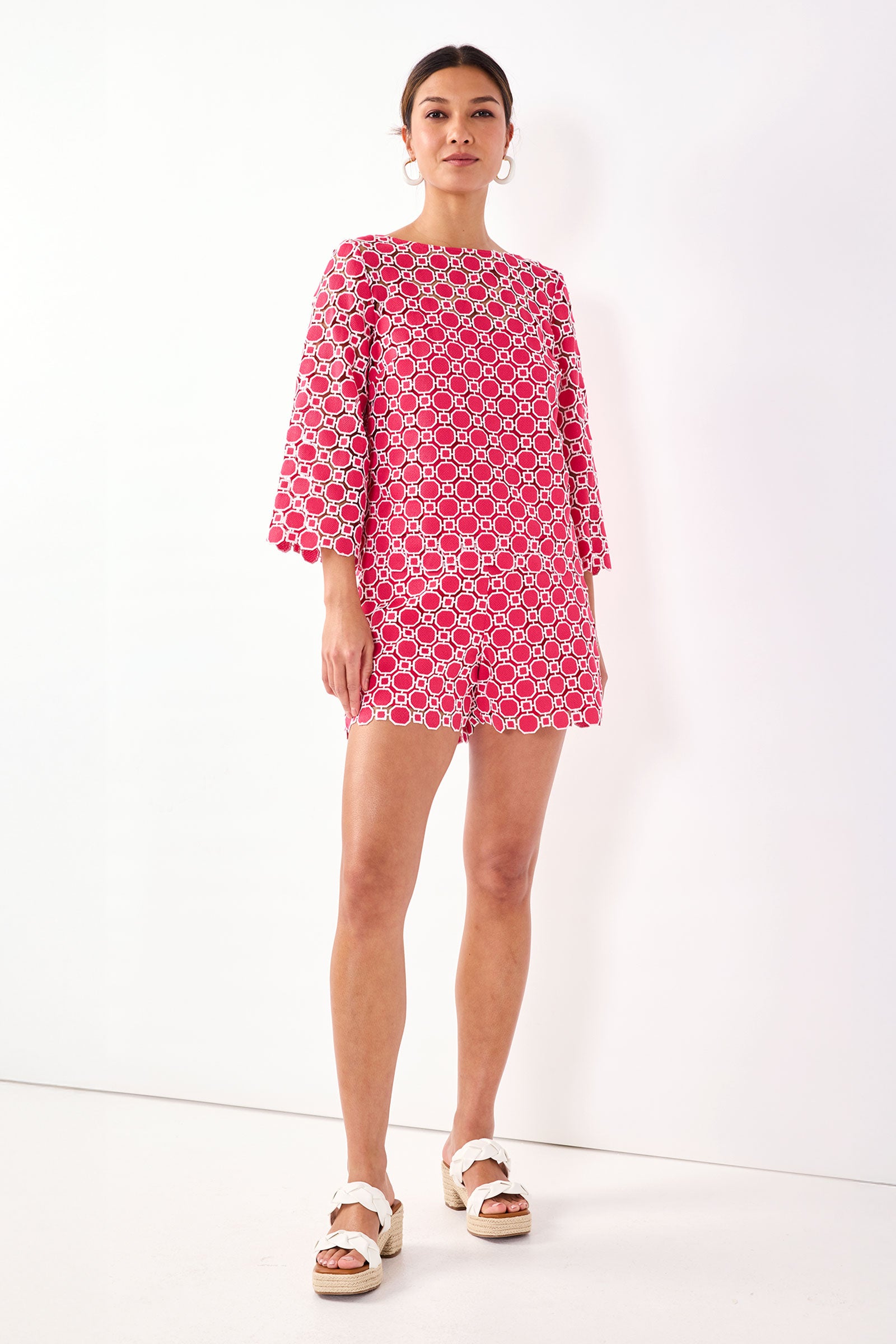 A woman stands against a white background wearing the Brightness Geo Lace Shorts in Cannes Coral with a matching geometric-patterned top, wide sleeves, and white platform sandals.
