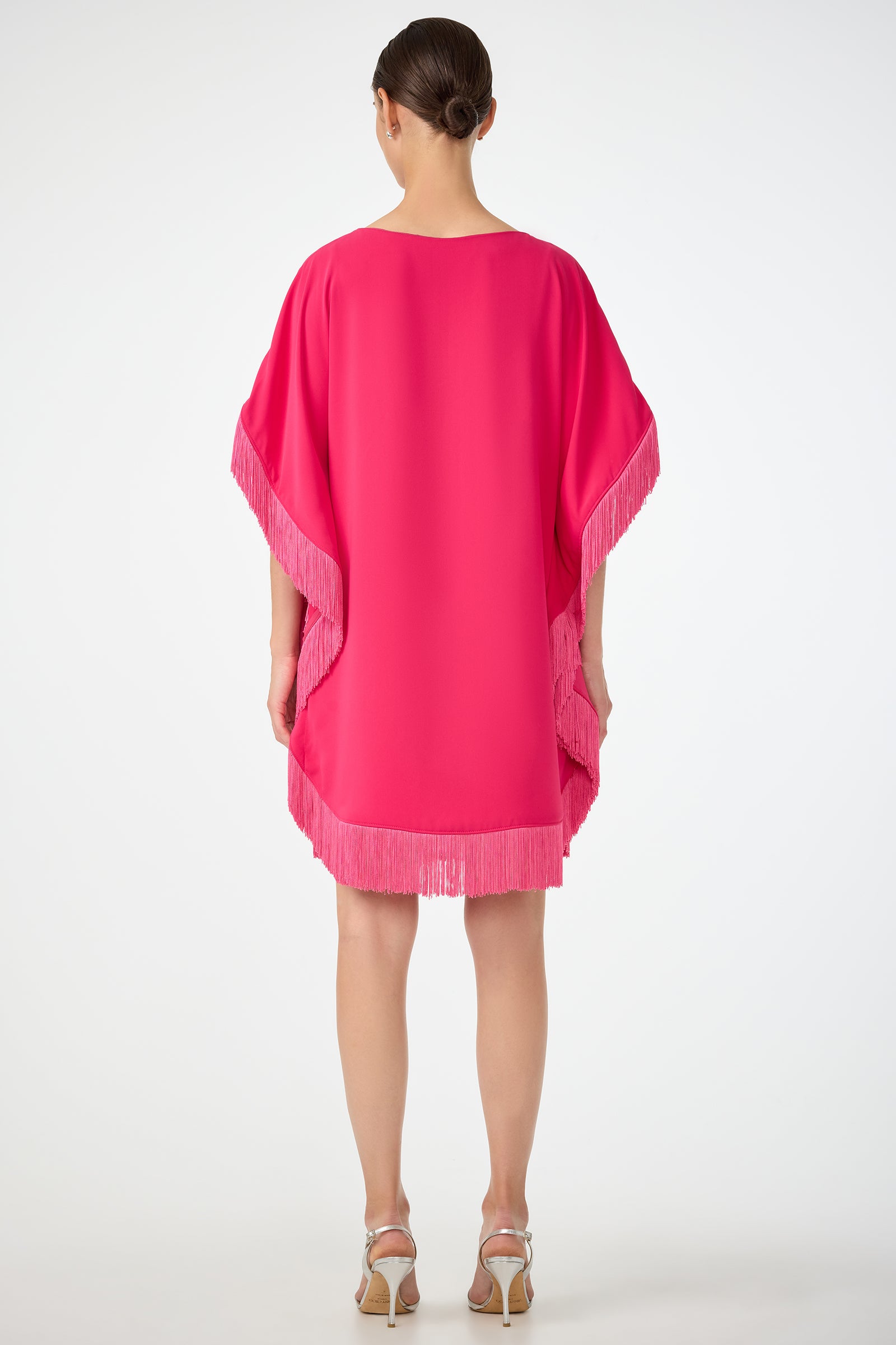 A woman faces away, wearing the Mini Neena Fringe Caftan Dress in bright pink with fringe trim on the sleeves and hem, styled with a low bun and white heeled sandals against a plain white background.
