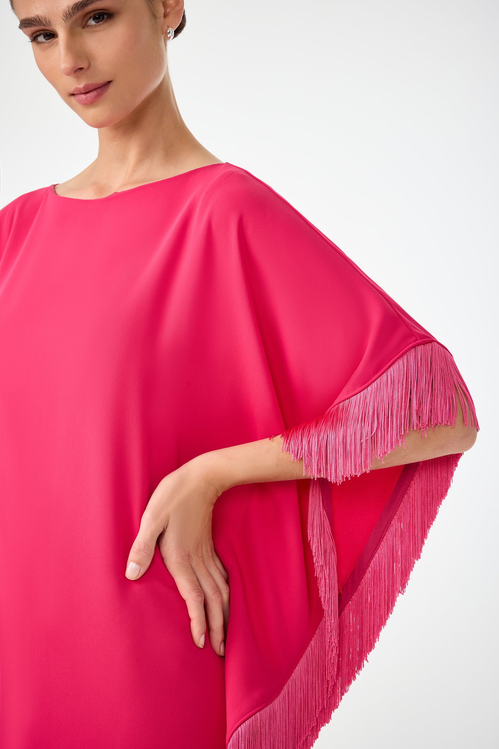 A woman poses against a plain white background wearing the Mini Neena Fringe Caftan Dress, a bright pink design with loose, flowing sleeves and matching pink fringe trim.