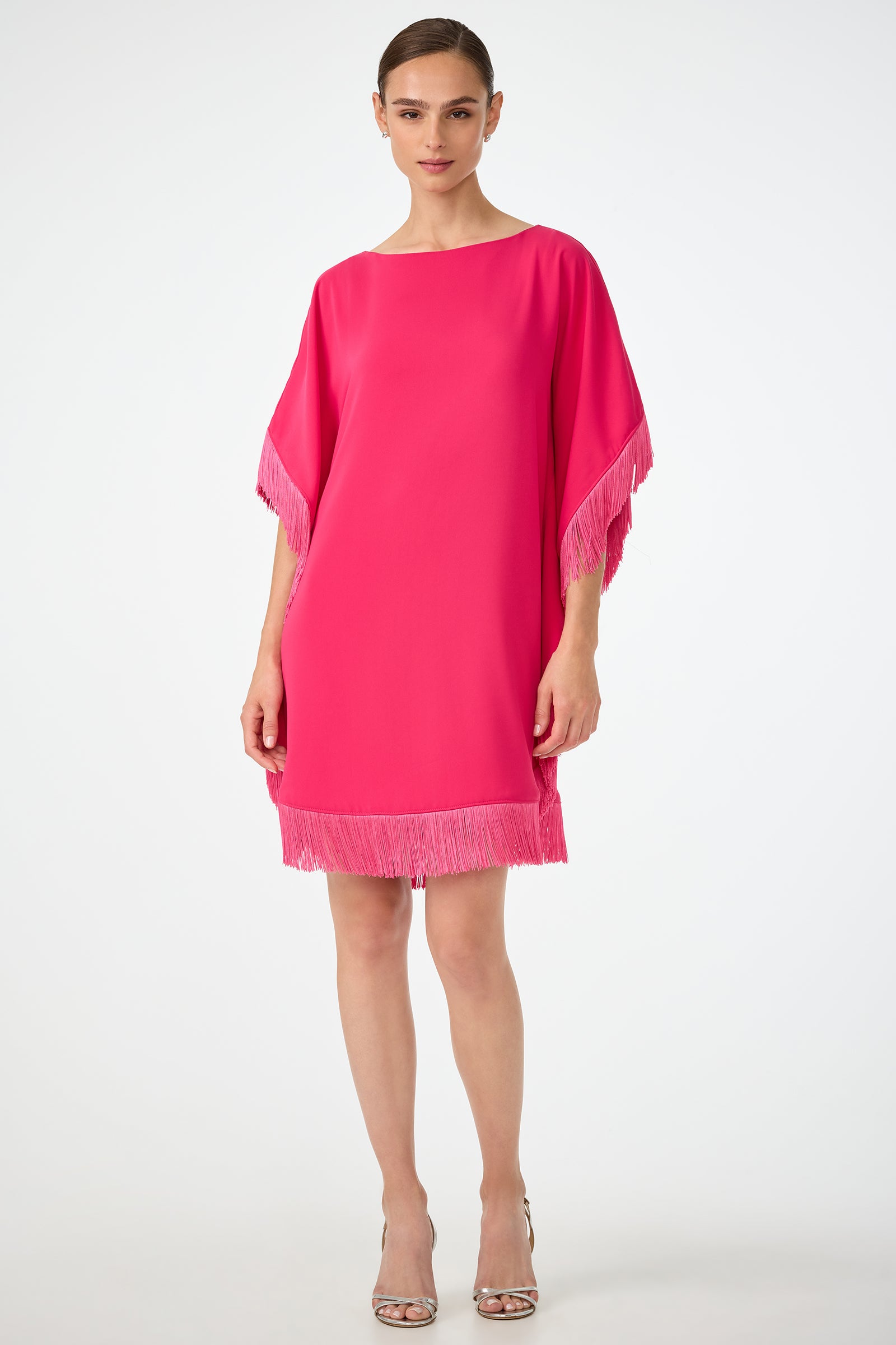 A woman stands against a plain background wearing the Mini Neena Fringe Caftan Dress in Forrest Rose, featuring fringe details on the sleeves and hem, paired with silver open-toe heels.