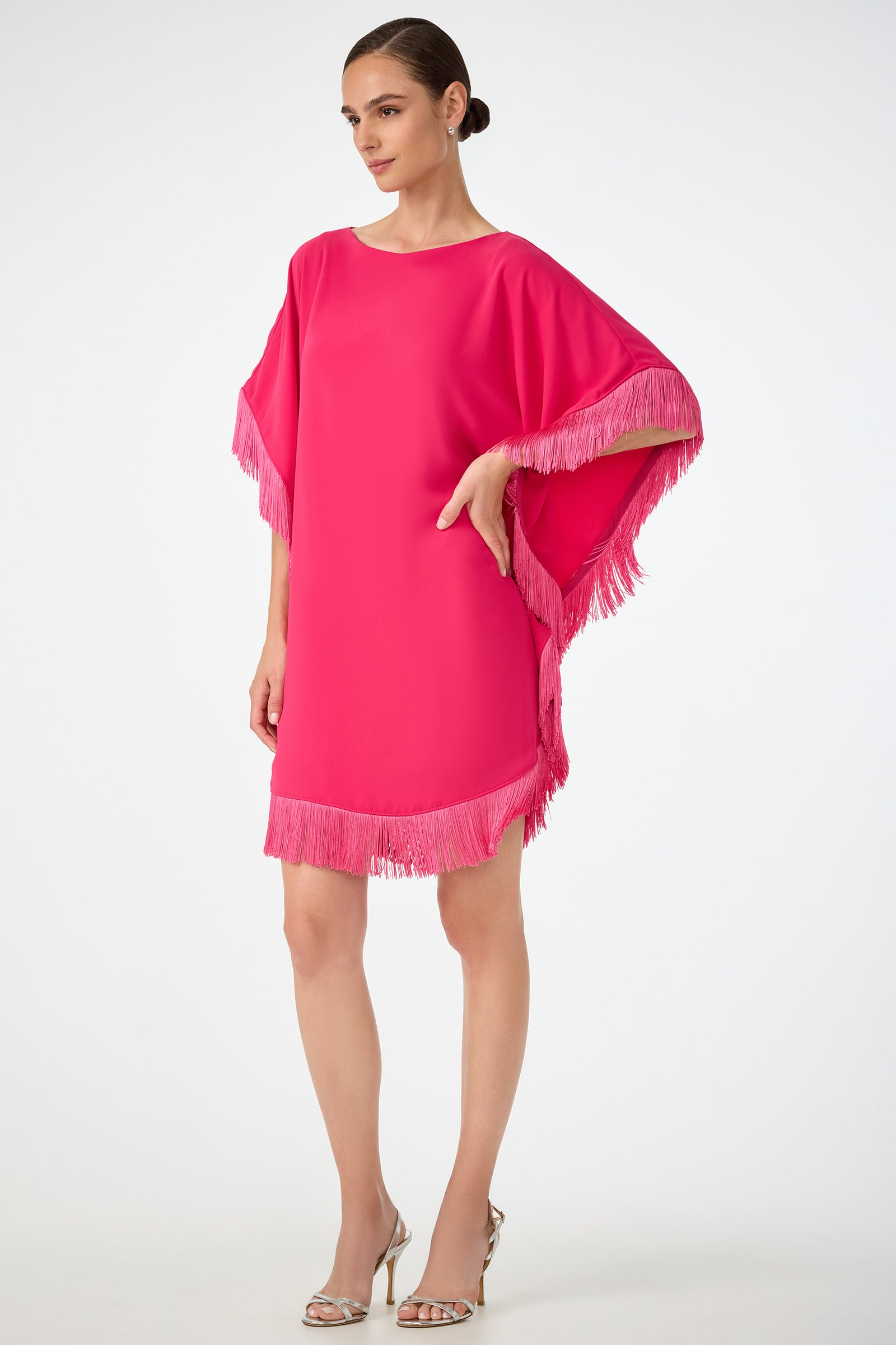 A woman wears the Mini Neena Fringe Caftan Dress, a bright pink, loose-fit style with fringe trim on the sleeves and hem, paired with silver open-toe heels. She stands against a plain white background.