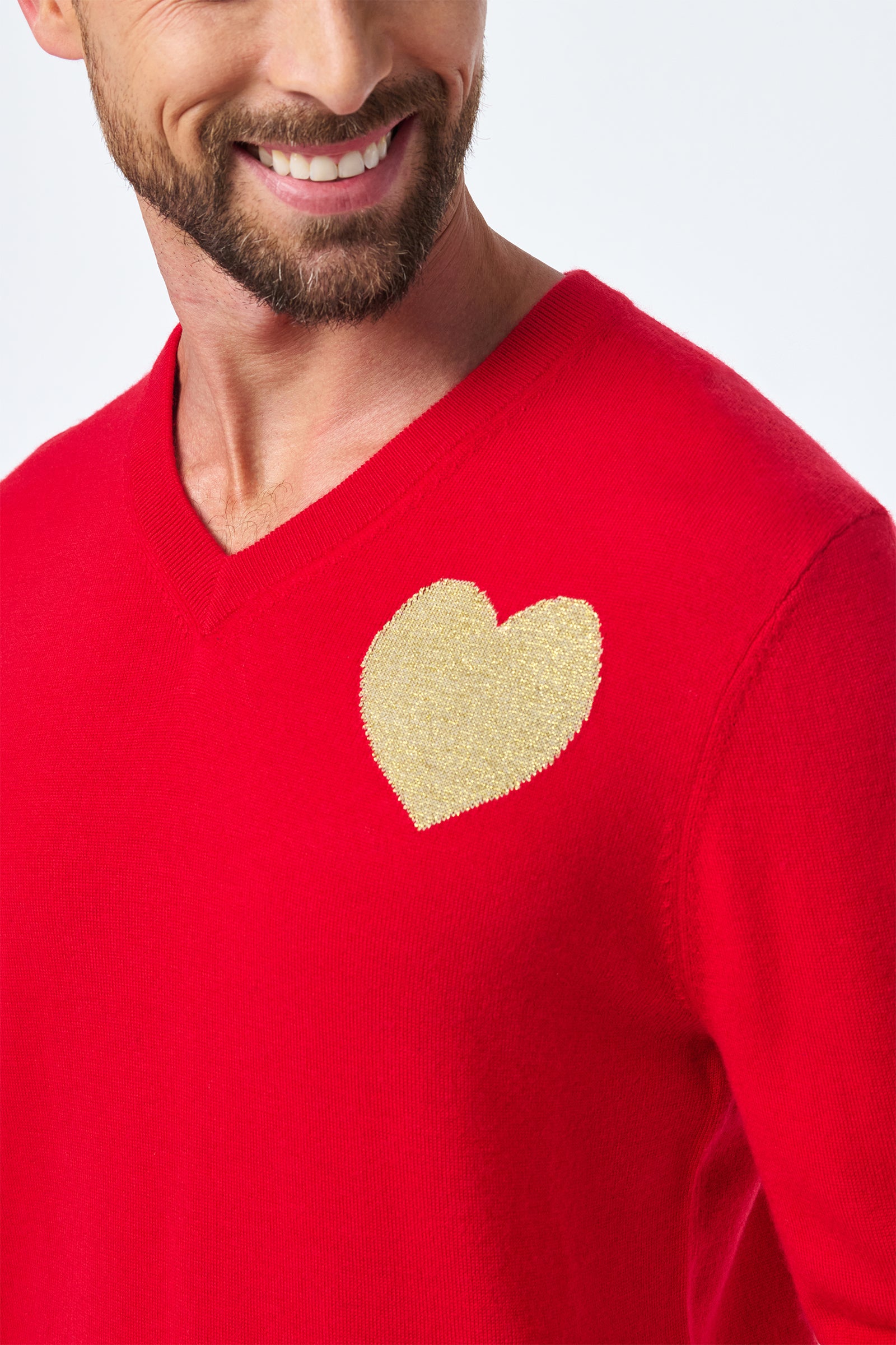 A smiling man wears the Lover Crewneck, a bright red sweater featuring a large gold heart patch on the upper left chest.