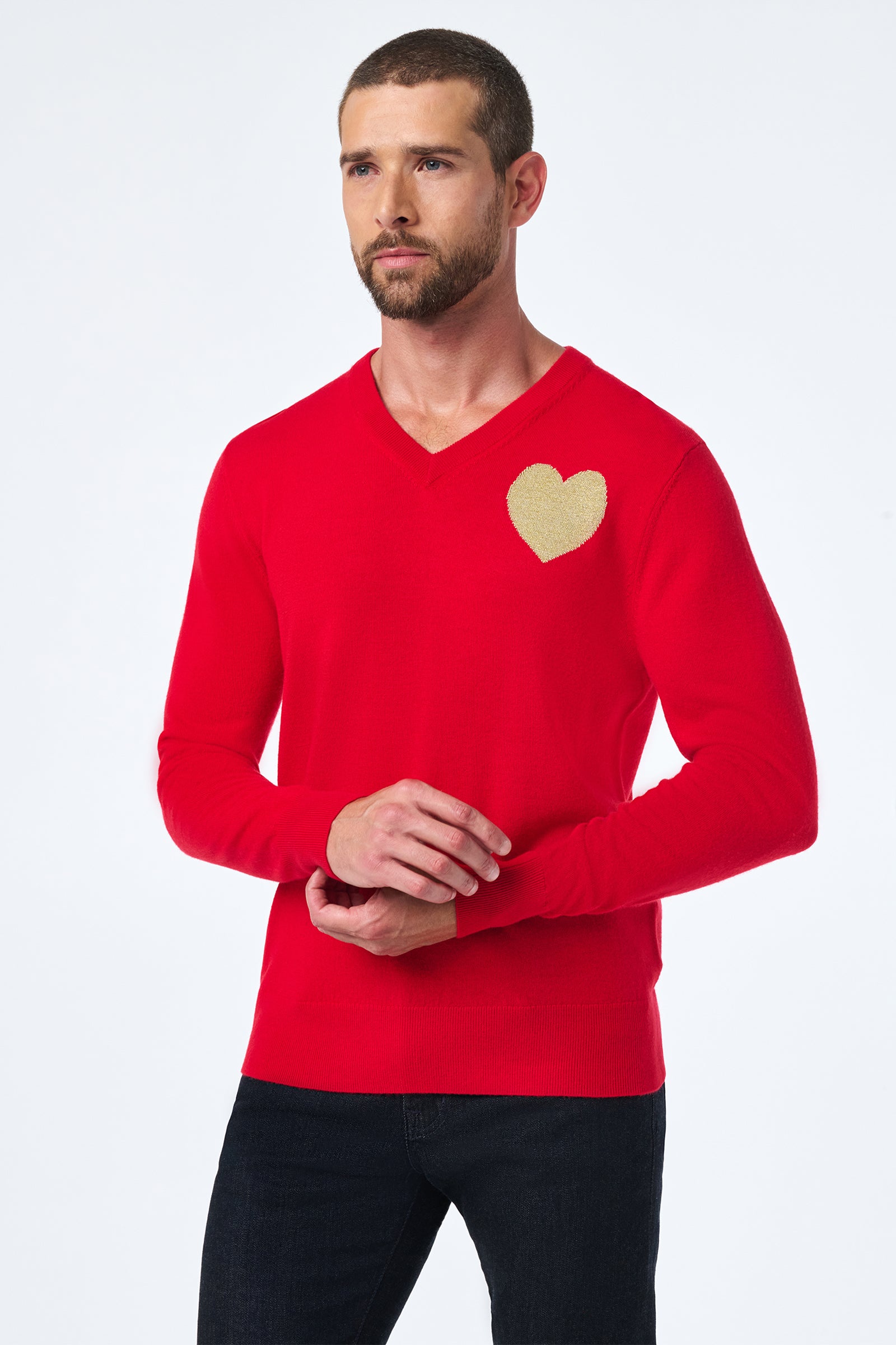 A man with short hair and a beard wears the Lover Crewneck, a bright red sweater featuring a gold heart patch on the left chest. He stands against a plain white background, looking slightly to the side.