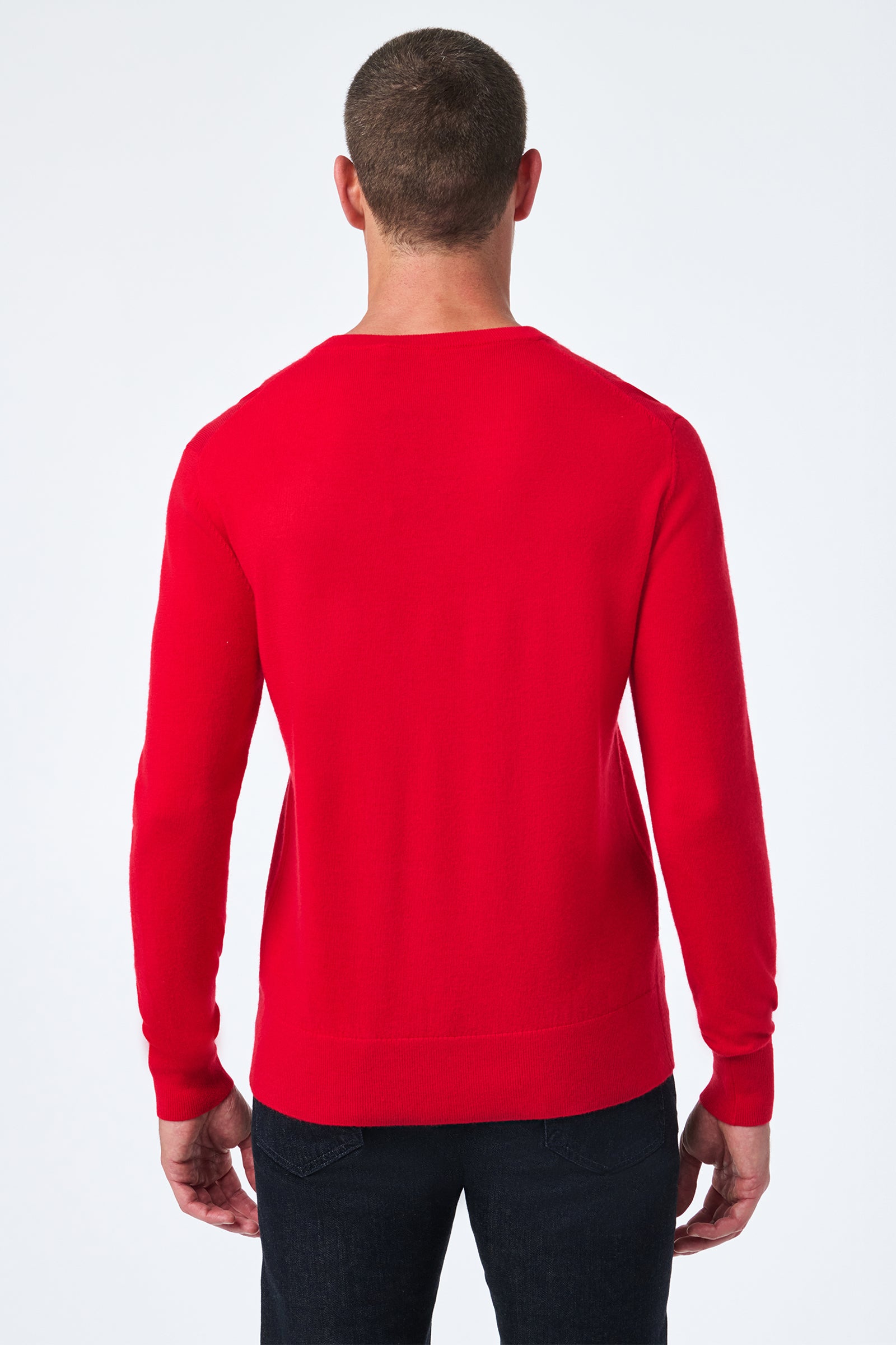 A man stands with his back to the camera, wearing the Lover Crewneck—a bright red long-sleeve sweater—paired with dark jeans against a plain white background.